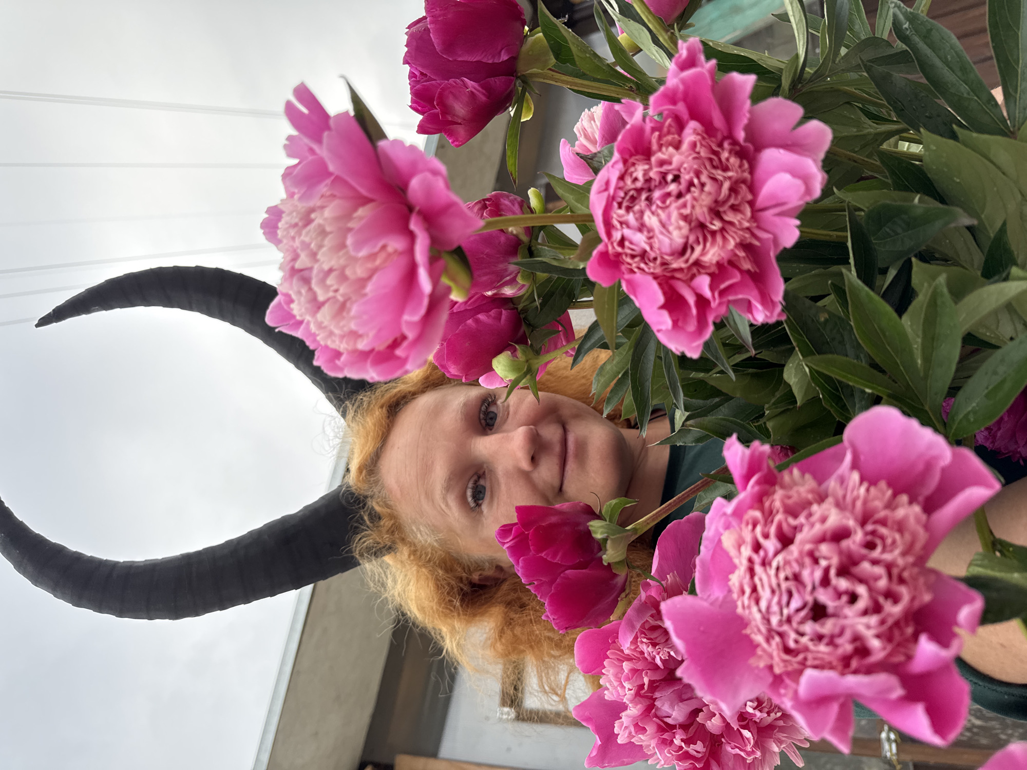 A close-up shot features a woman with curly ginger hair and pale blue eyes, wearing black devil horns and holding a large bouquet of bright pink peonies. The woman is looking towards the camera with a slight smile. The background is blurred, showing hints of a structure that could be a building or fence, and an overcast sky. The image appears to be taken outdoors, possibly in a garden or natural setting within the city of Chaikovsky, Russia.