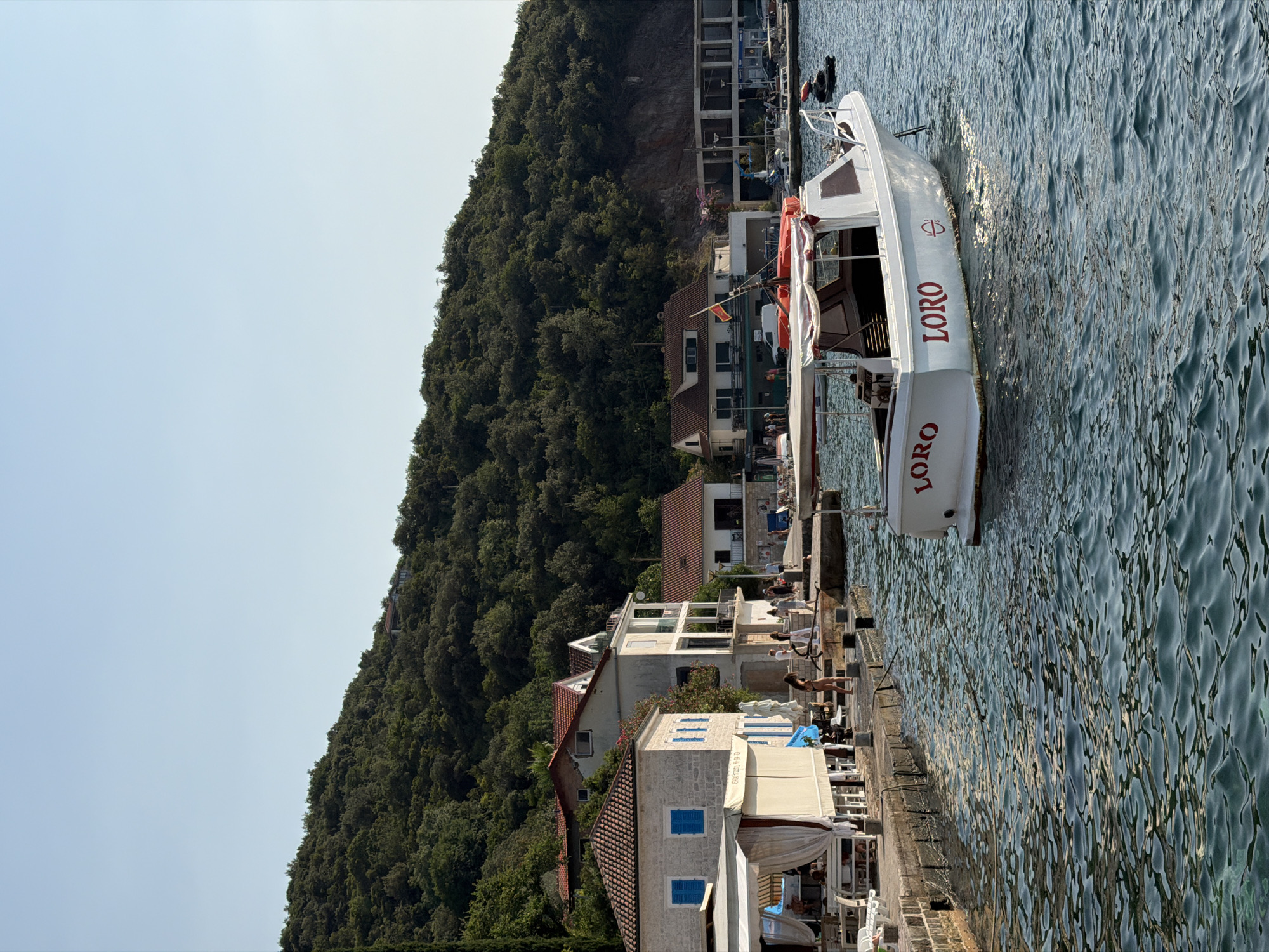The image depicts a sunny day in Zelenika, Montenegro, with a focus on the waterfront and the adjacent buildings. A white motorboat, with "LORO" emblazoned on its hull in red letters, is anchored in the foreground, bobbing gently on the rippling blue water. The boat has a canopy providing shade over part of its interior.
To the left of the boat, a stone pier extends outwards. Along the pier, there are several buildings. The closest building on the pier is a light-colored structure with a white canopy extending over a seating area, suggesting an outdoor cafe or restaurant. Behind this, a two-story building with traditional red-tiled roofs and light-colored facades is visible, featuring blue shutters on some windows. Further down the pier, more buildings with red roofs and contemporary architectural elements are present.
Several people are scattered along the pier, some walking, others appearing to be seated at tables or by the water. A few individuals are wearing swimwear. A jet ski is visible in the water behind the "LORO" boat.
The background is dominated by a lush green, tree-covered hillside that rises steeply behind the buildings. The sky is a clear, pale blue, indicating fair weather. The overall scene conveys a relaxed, coastal atmosphere.