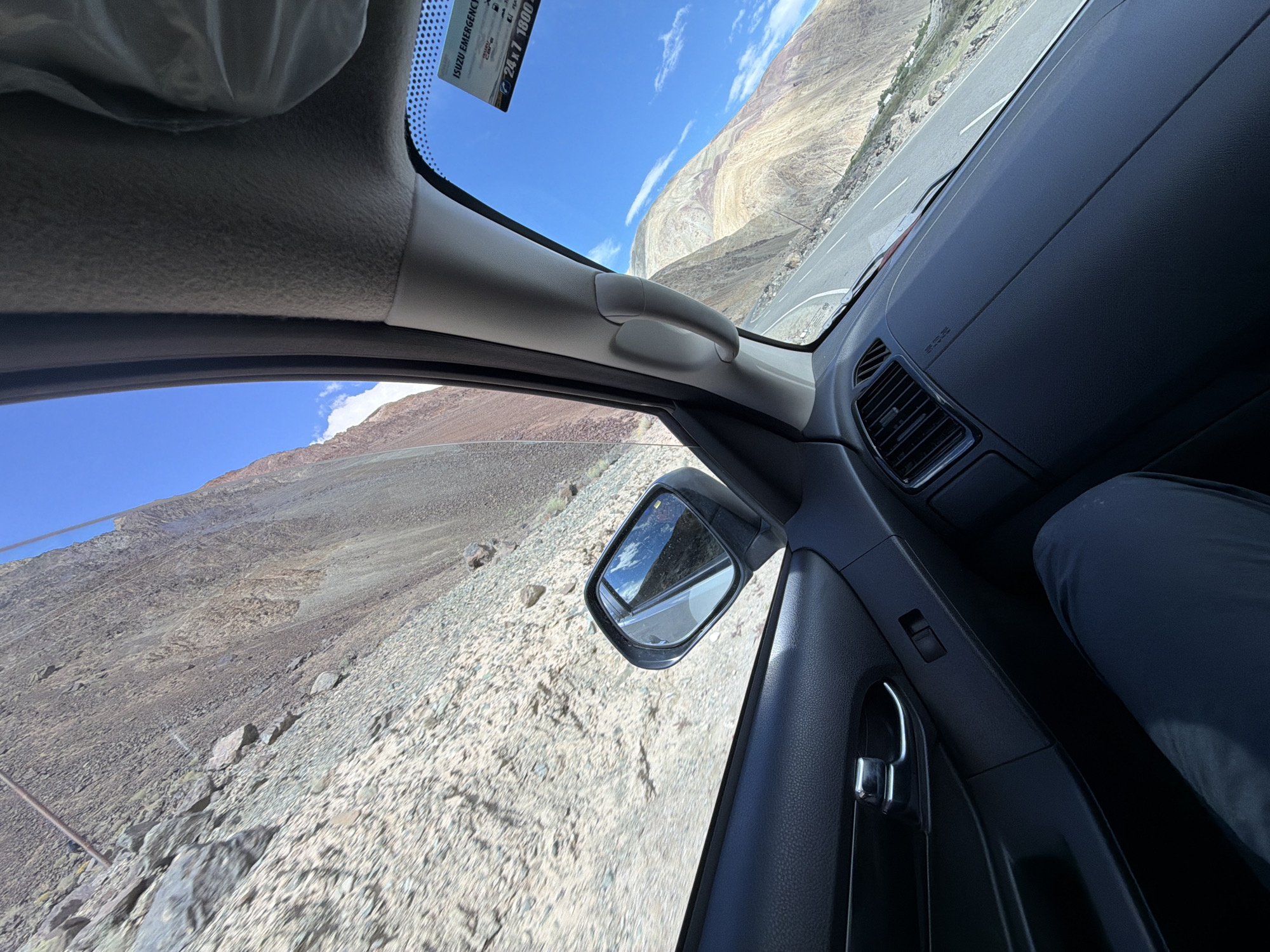 The image shows a view from inside a car, looking out the passenger side window and through the windshield. The car is on a paved road that winds through a mountainous, arid landscape with rocky slopes and sparse vegetation. The sky is clear blue with a few wispy clouds. A side-view mirror reflects the road and mountains. On the dashboard, an "ISUZU EMERGENCY" sticker is visible. A person's leg, clad in dark pants, is visible in the lower left corner of the frame. The environment appears to be the city of Chumathang, India, known for its hot springs and proximity to the Himalayas.