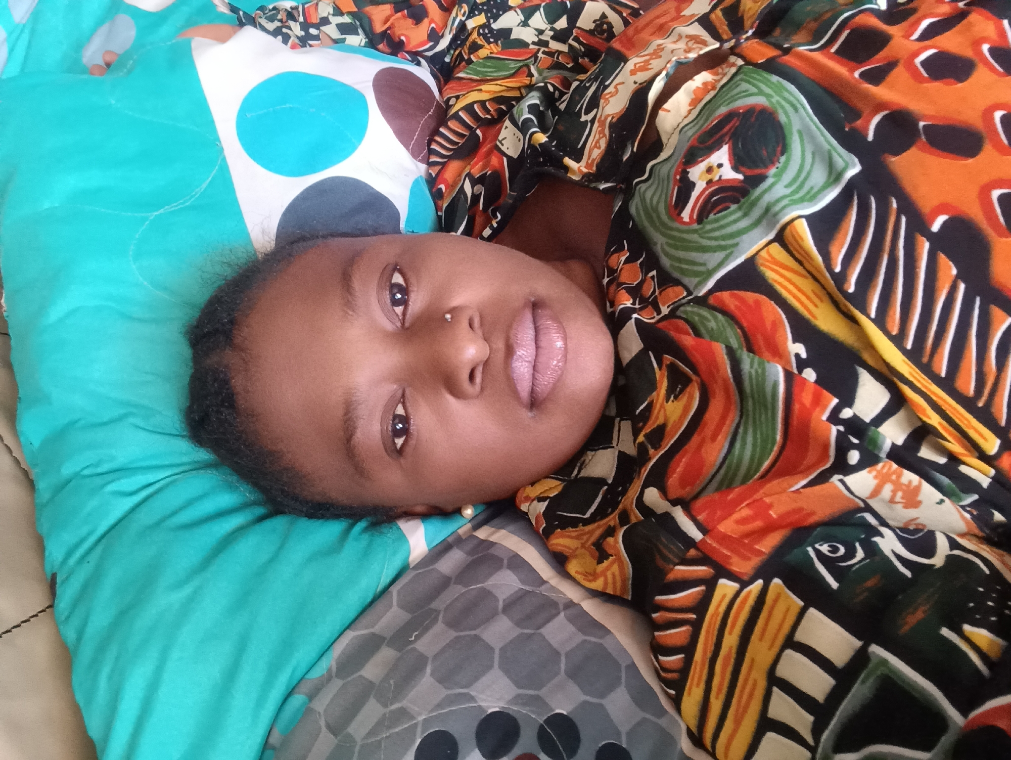 A young person with dark hair is lying down on a bed. They are resting their head on a blue and white polka-dot pillow. The individual is covered with a colorful, patterned fabric. The background includes a grey patterned sheet and hints of a light brown surface. There is no specific visual information to place this image definitively in Jos, Nigeria. Therefore, the precise location cannot be verified from the image alone.