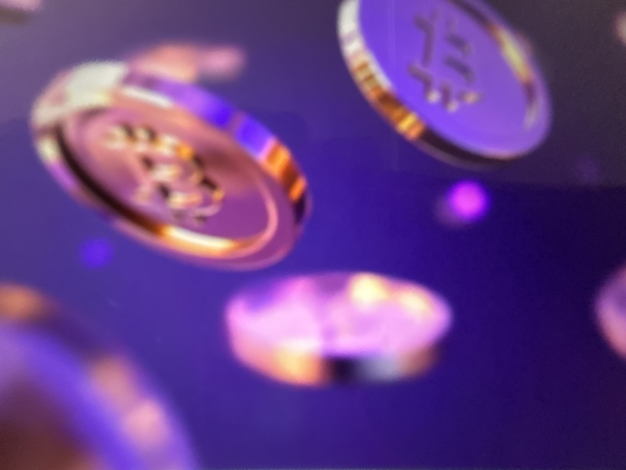 The image depicts a dark purple background with several out-of-focus, circular, coin-like objects that appear to be floating. The objects have a metallic sheen and reflect light, showing streaks of pink, orange, and white. One coin in the upper right and another in the lower right are more distinctly visible, appearing to have etched designs on their surfaces, though the details are obscured by blur. There are also blurry areas of pink and purple light scattered throughout the background. The scene is abstract and appears to be digitally rendered or a heavily edited photograph. There is no discernible connection to the city of Gelendzhik, Russia, or any real-world environment.