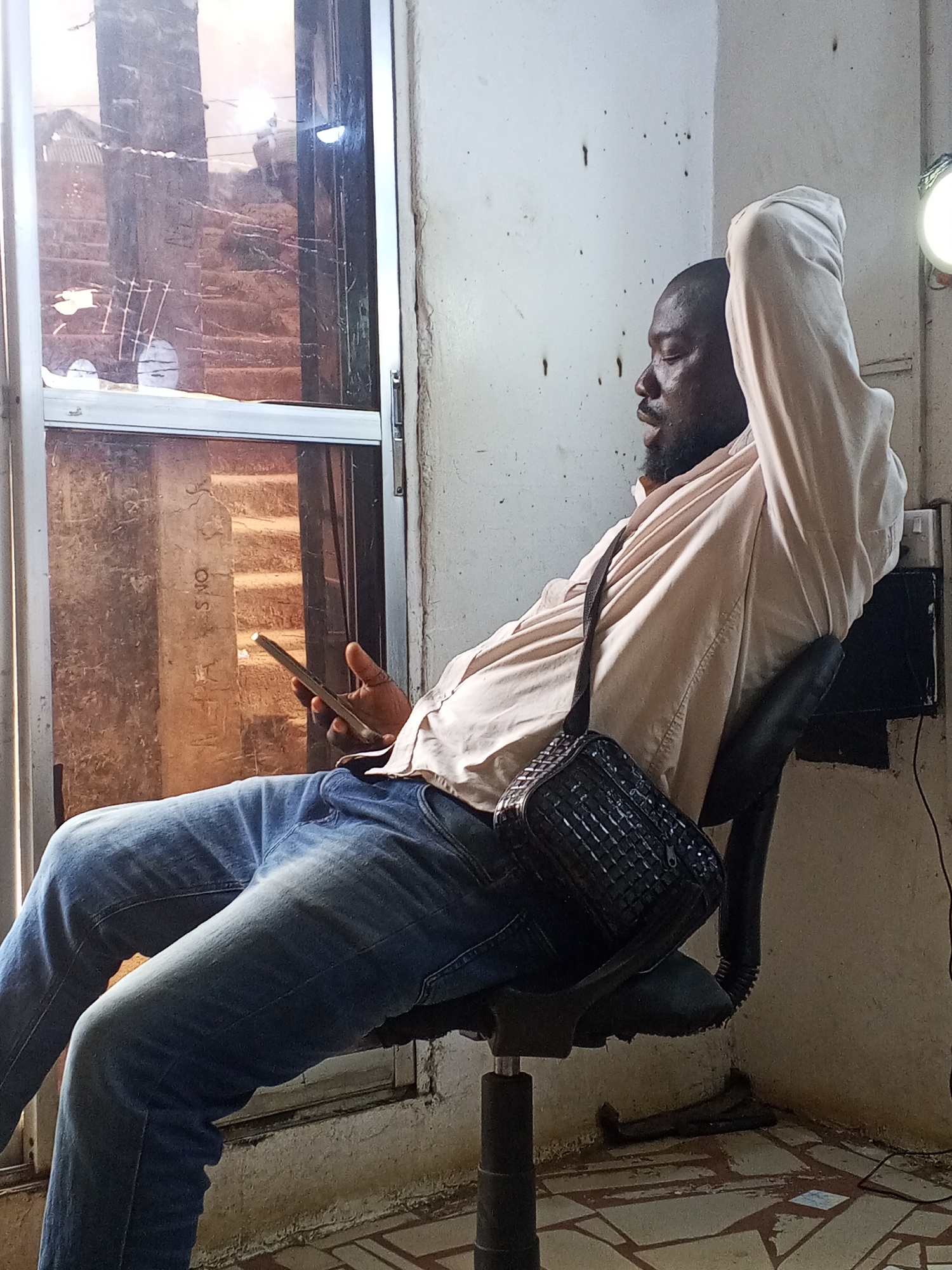 A man is sitting on an office chair next to a window. The man is Black, and he has a beard. He is wearing blue jeans, a light pink shirt, and a black crossbody bag. He is holding a cell phone in his left hand and resting his right arm above his head. The window has a view of a building across the street. The wall behind the man is white, and there are some dark stains on it. There is a light fixture on the wall to the right of the man. The location of the image is the city of Unknown, Unknown.