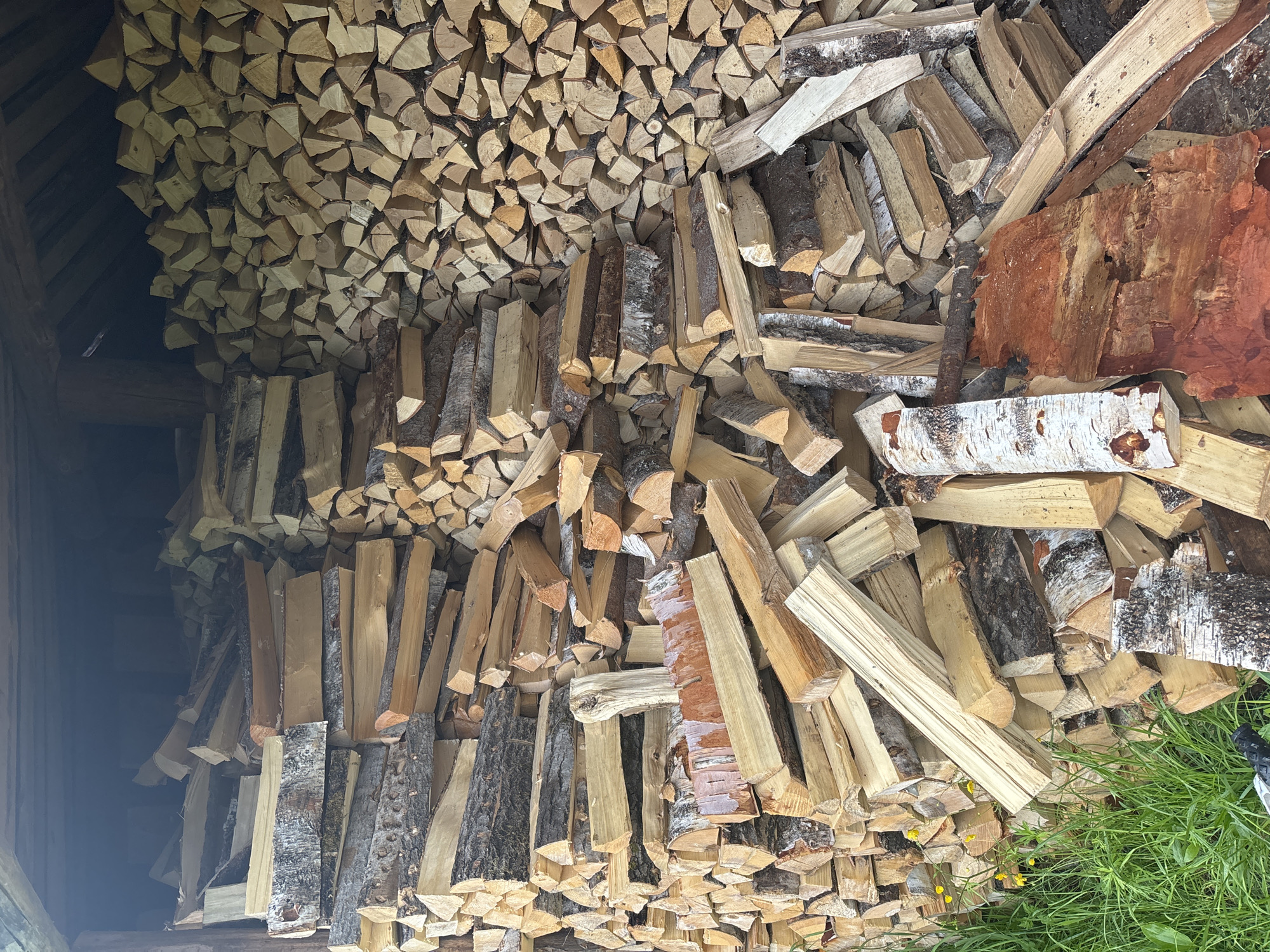 A vertical, full-shot photograph shows a large stack of chopped firewood filling a rustic, open-sided wooden structure. The wood is piled high, with some pieces showing bark and others exhibiting the light-colored wood grain. The firewood is arranged in a somewhat orderly fashion, but there are also loose pieces and debris scattered throughout. The background of the structure is dark, suggesting a covered or shaded area. In the foreground, to the left of the firewood, there is green grass with a few small yellow wildflowers. The overall scene appears to be an outdoor storage area for firewood, possibly in a rural or semi-rural setting. The image does not contain any people, nor does it provide specific details that would identify its location in the city of Курган, Russia.