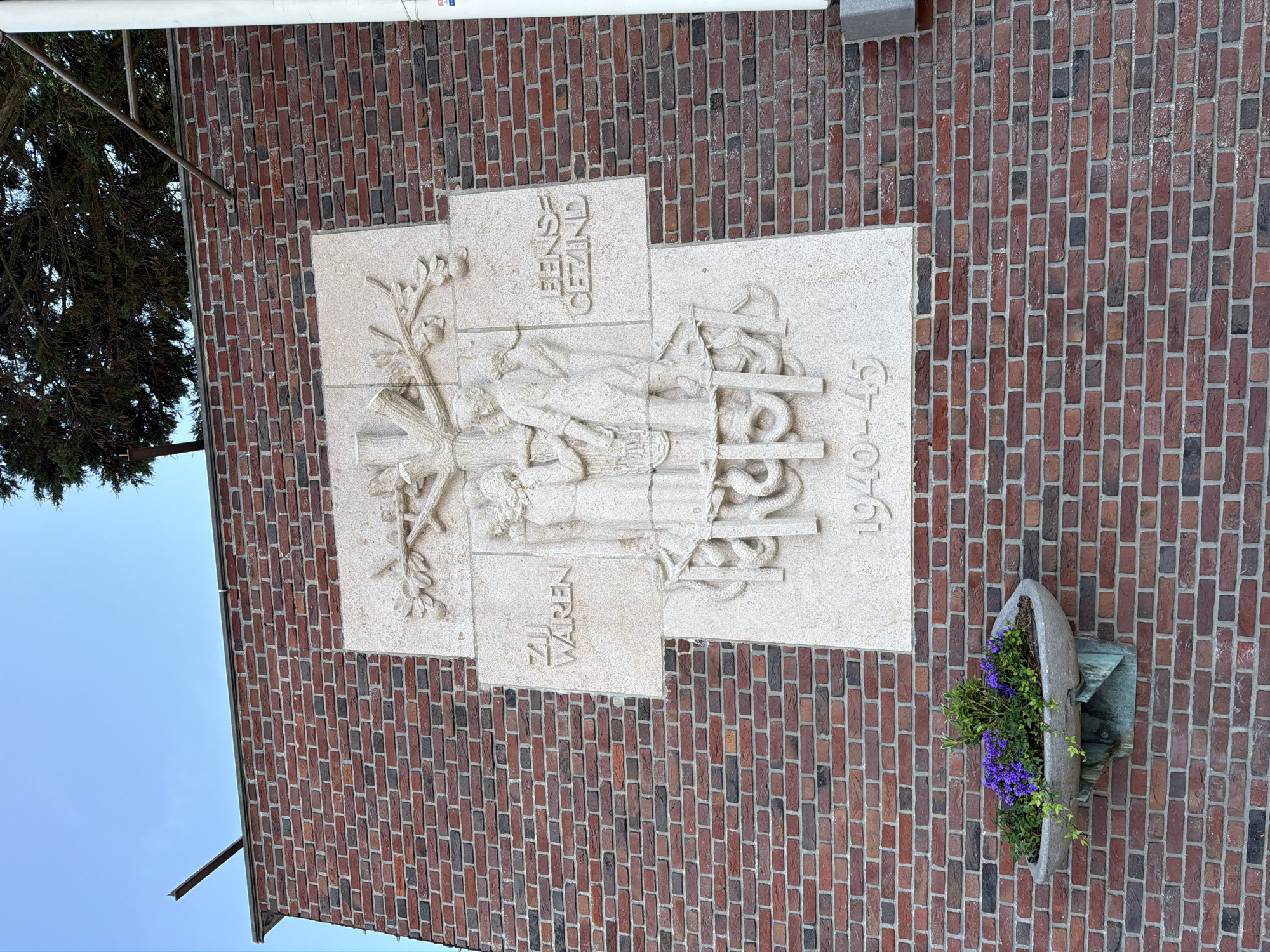 The image shows a relief sculpture on a brick building in The Hague, Netherlands. The sculpture depicts a tree with branches and fruit, with three figures standing beneath it. The figures appear to be a family, with a man and woman holding hands and a child standing between them. The sculpture is made of stone and is inscribed with text that reads "ZIJ WAREN EENS GEZIND" and "1940-45". The text translates to "They were once united" and the dates refer to the period of World War II. The sculpture is located on the facade of a building, with a flower planter on the lower left side. The sky is visible in the background, with some tree branches hanging over the building. The building is constructed of red brick.