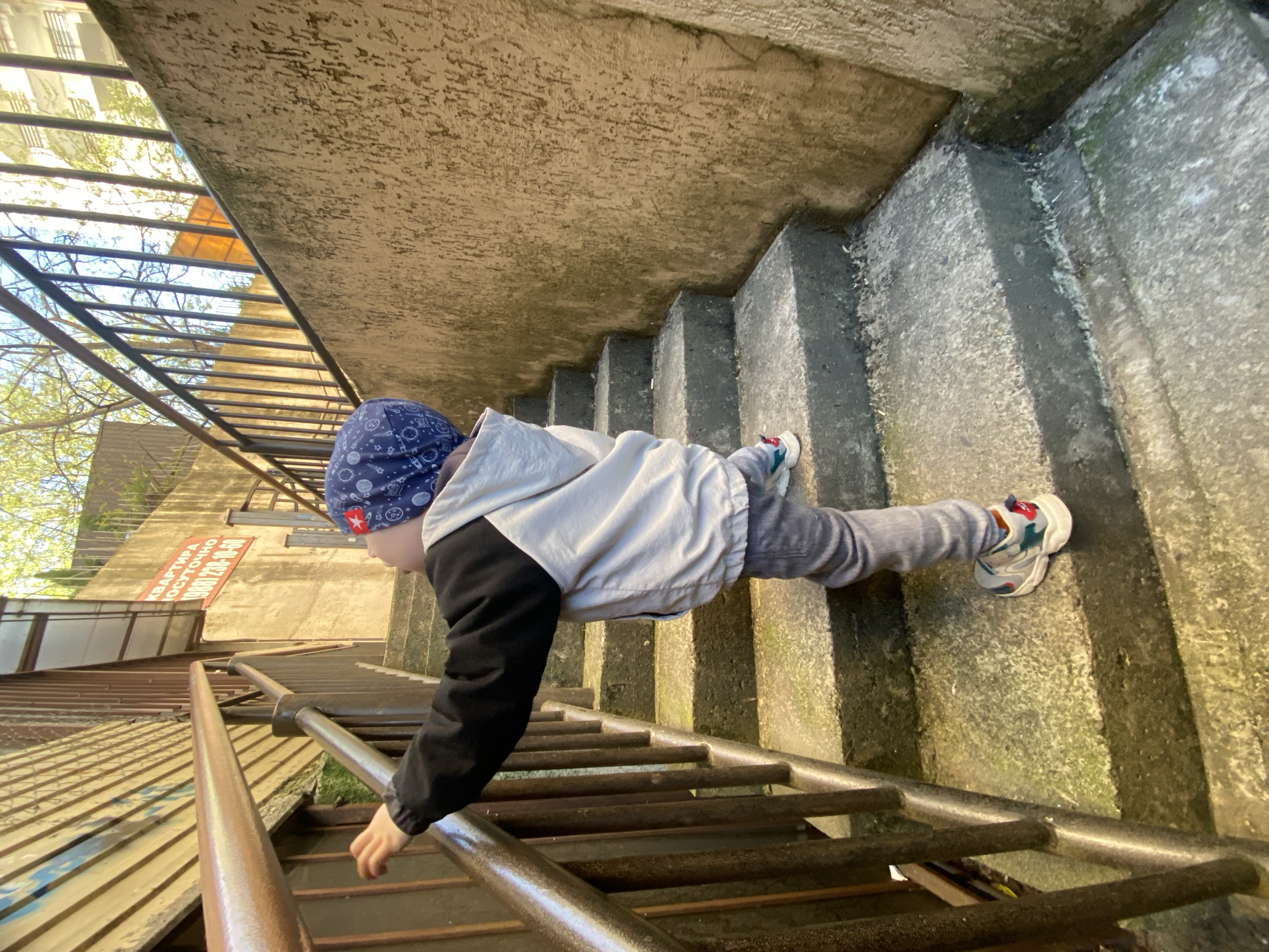 An outdoor scene captured from a slightly elevated perspective shows a young child carefully descending a flight of concrete stairs. The child, dressed in a blue beanie with white patterns, a light grey jacket with black sleeves, grey pants, and colorful sneakers, is holding onto a robust metal railing with their right hand for support. The railing consists of multiple horizontal metal bars.
The concrete stairs appear old and weathered, with visible dirt, grime, and possibly moss accumulating on their surfaces and edges. Bright natural light illuminates the scene, casting shadows and suggesting a sunny day, likely morning or afternoon.
In the background, to the left of the child and railing, there are green trees with lush leaves and what appears to be a metal grate structure or fencing. Further back, a building or wall is visible, on which a reddish-orange sign is mounted. The sign displays Russian text: "КВАРТИРА ПОСУТОЧНО" (Apartment for daily rent) and a phone number "(988) 230-10-60". The setting suggests an urban or semi-urban pathway or alleyway, providing access between different levels, located in Sochi, Russia. The child's posture indicates focused, careful movement down the steps.