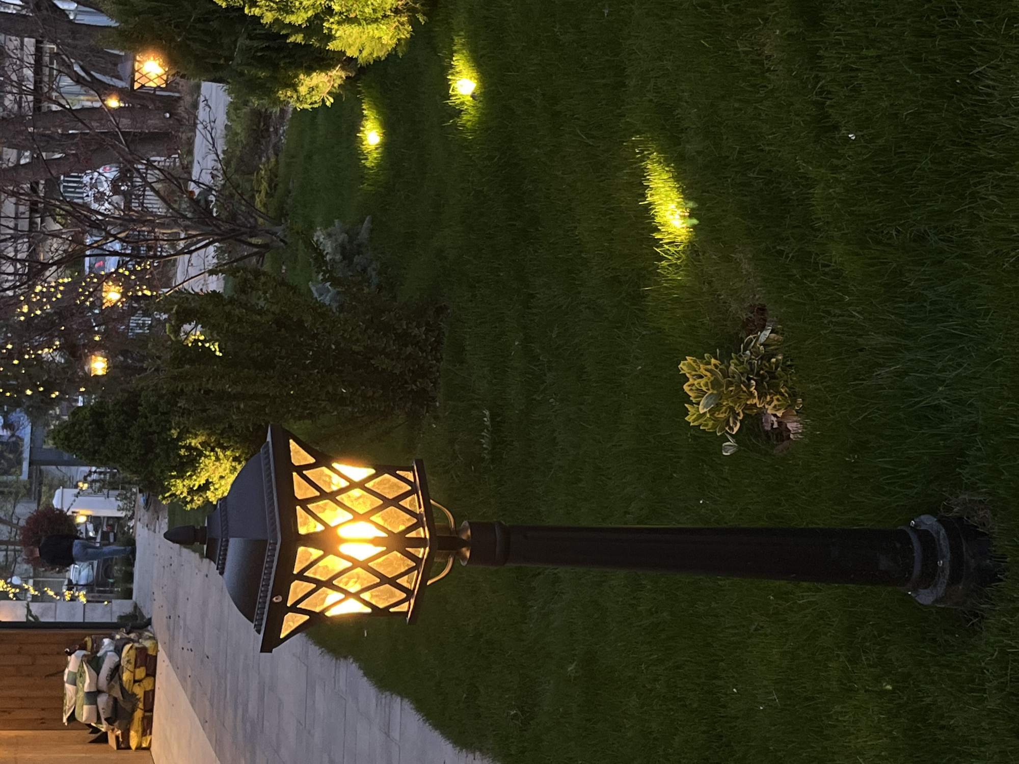 This image depicts an outdoor scene at night, likely a garden, park, or restaurant patio. The setting is illuminated primarily by artificial lights, creating a warm and inviting atmosphere.
In the foreground and central to the image is a tall, black ornamental lamppost. Its lantern emits a bright, warm yellow-orange light through a diamond-patterned enclosure. To the left of the lamppost, a light-colored paved path stretches into the distance, guiding the viewer's eye.
The area around the path and the lamppost is covered in lush, dark green grass. Several smaller, bright yellow spotlights are embedded within the grass, casting distinct beams of light that highlight various patches of turf. Further into the background, scattered among trees and bushes, are more warm-toned lights, some appearing as small string lights draped over branches. The foliage is generally dark, indicating the nighttime setting, though some greener, possibly coniferous, trees are visible on the right. Some trees in the mid-ground exhibit bare branches.
In the far background, several indistinct figures can be seen moving or standing near what appears to be an outdoor seating area or building. One person is more clearly visible walking on the paved path. On the very left side of the frame, near the path, there is a stack of large sacks or bags, possibly containing sand or other materials, with visible yellow, white, and green patterns.
The time of day is clearly evening or night, characterized by the darkness and the reliance on artificial illumination. There is no visible text in the image. The location is Sofia, Bulgaria.