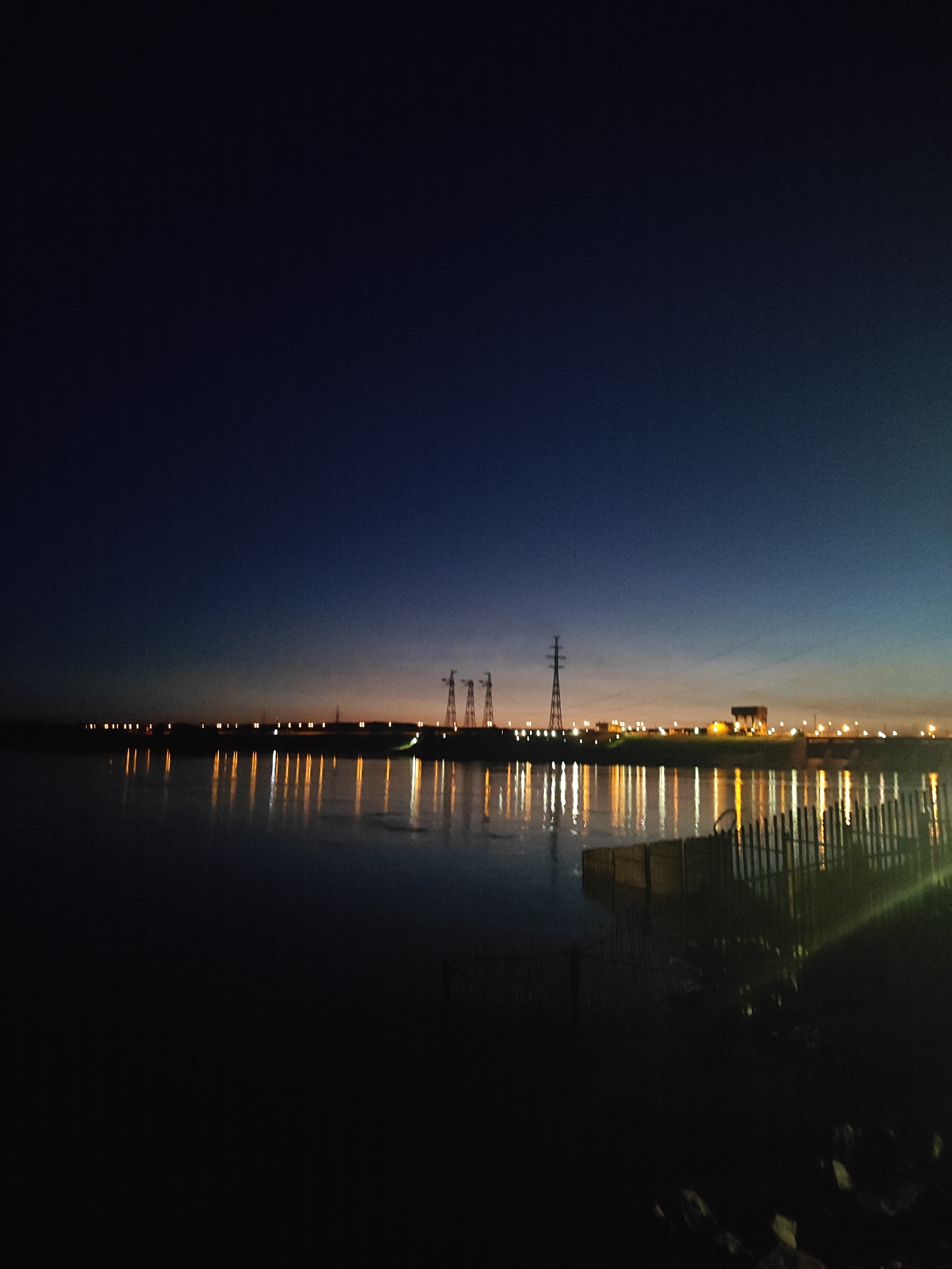 This nocturnal landscape captures a wide view of a body of water, likely the Kama River or a reservoir near Chaikovsky, Russia, at twilight or night. The sky transitions from a deep indigo at the zenith to a lighter blue, with a warm orange and yellow glow visible just above the distant horizon.
Across the water, a long line of artificial lights illuminates the far bank, casting bright, vertical reflections onto the calm surface. These lights suggest a populated area or industrial infrastructure. Silhouetted against the glowing horizon are several tall, lattice-structured electricity pylons, characteristic of power transmission. To the right of these pylons, a large, box-like building or structure is also visible, likely part of a hydroelectric station or a related industrial complex, aligning with the location near the Votkinsk Hydroelectric Station.
In the immediate foreground on the right, the shadowy outline of what appears to be a wooden fence or barrier extends into the water, suggesting a dock or embankment. The water itself is dark, with the prominent reflections being the main source of light. No people or specific activities are discernible, and no text is visible within the image. The scene conveys a sense of quiet solitude with a strong human presence indicated by the distant lights and structures.