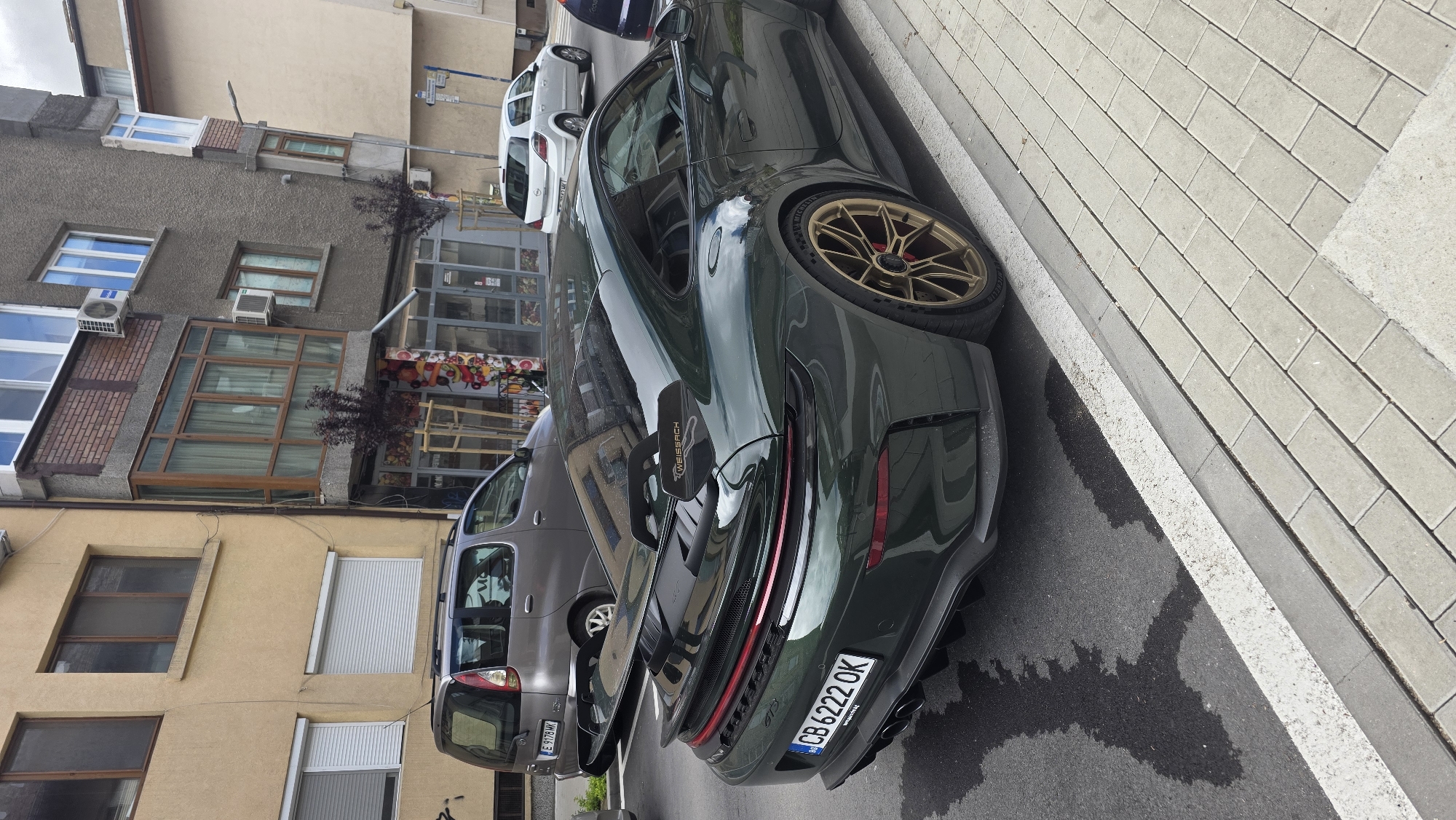 The image shows a green Porsche 911 GT3 parked on a street in Blagoevgrad, Bulgaria. The car has gold wheels and a "Weissach" package rear wing. The license plate reads "CB 6222 OK". The car is parked next to a gray minivan. Behind the minivan are buildings with various windows and storefronts, and a sidewalk paved with grey bricks. The pavement near the car is wet.