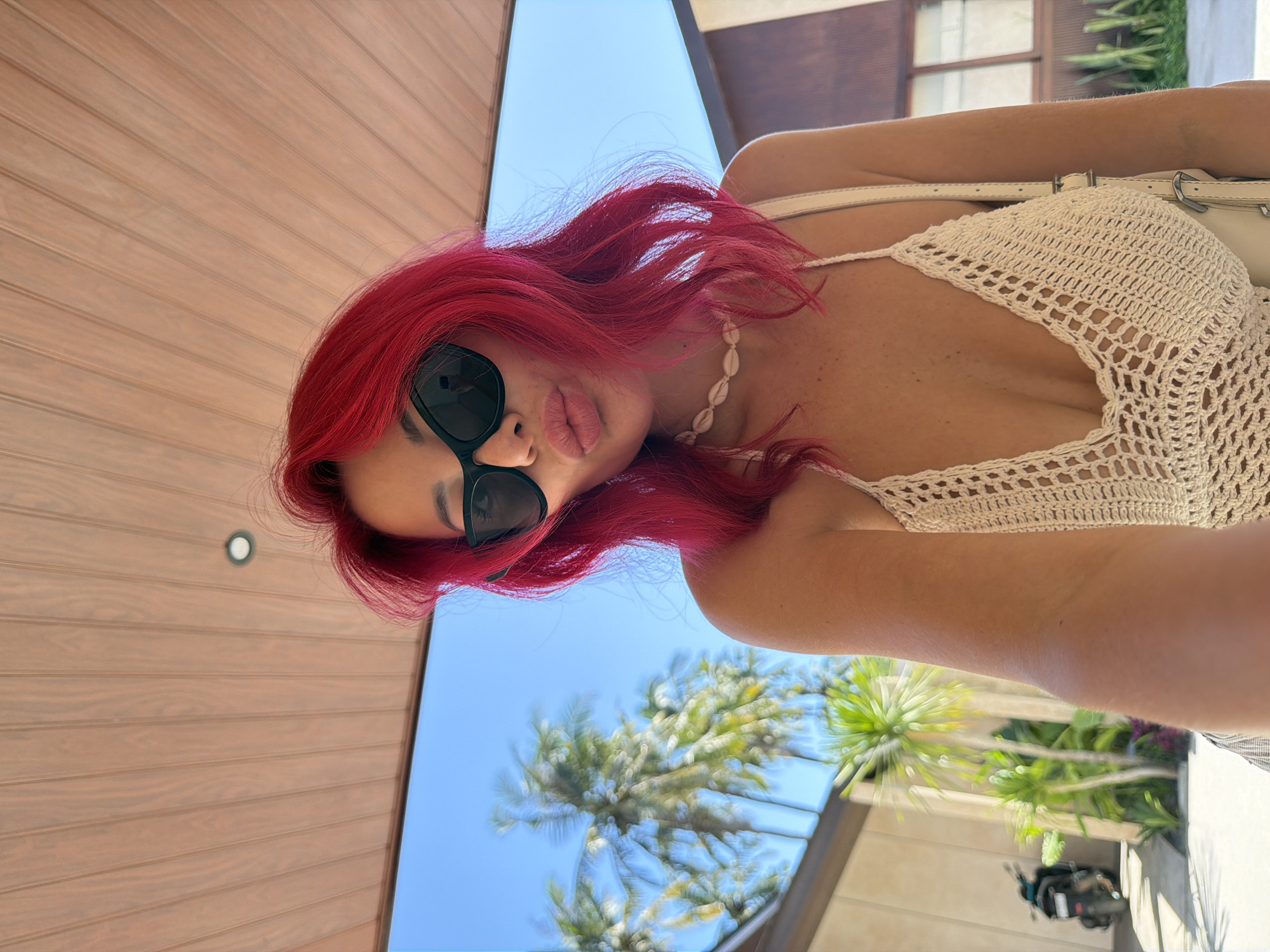 A woman with bright pink hair and sunglasses is taking a selfie outdoors. She is wearing a white crocheted top and a shell necklace. The background shows palm trees and a building, suggesting a tropical location. To the left, a scooter is parked on a paved area. The scene appears to be in Kuta, Indonesia, indicated by the presence of palm trees and Balinese-style architecture.