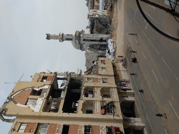 The image displays a scene of significant urban destruction in Khan Yunis, Palestinian Territory. A prominent multi-story beige building on the left exhibits severe structural damage, with large sections of its facade missing, exposing interiors across multiple floors. The upper levels show a partially destroyed arched architectural element, and balconies with white balustrades are visible, some with clothes hanging. To the right, a mosque with a white dome and a tall, slender minaret also shows extensive damage to its structure, standing amidst a broad area of rubble, sand, and debris that extends to its base. A paved street with faded white lane markings occupies the foreground. Several individuals, primarily appearing to be children, are present on the street: one person walks away on the far left, another crouches on the sidewalk, a small group plays with a soccer ball in the center, and an individual stands further right. Another person in reddish attire is visible standing in the sandy, rubble-filled area near the damaged mosque. The sky above is clear.