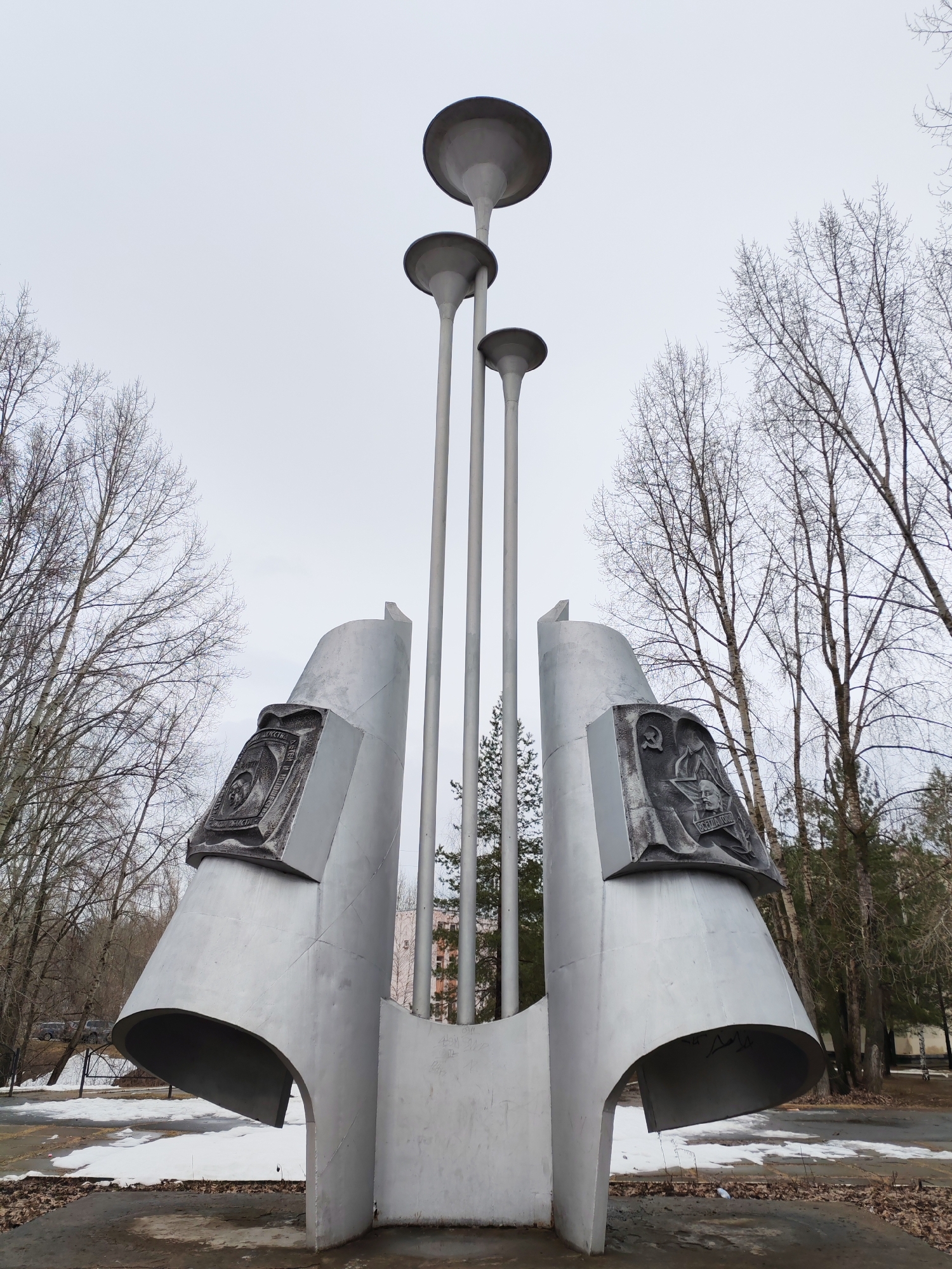 The image shows a metallic monument in Chaikovsky, Russia. The monument consists of two curved, flared structures with embedded plaques, flanking three tall, cylindrical poles topped with disc-shaped elements. The sky is overcast. Bare trees surround the monument. Patches of snow remain on the ground. The immediate foreground consists of a paved surface covered with debris.