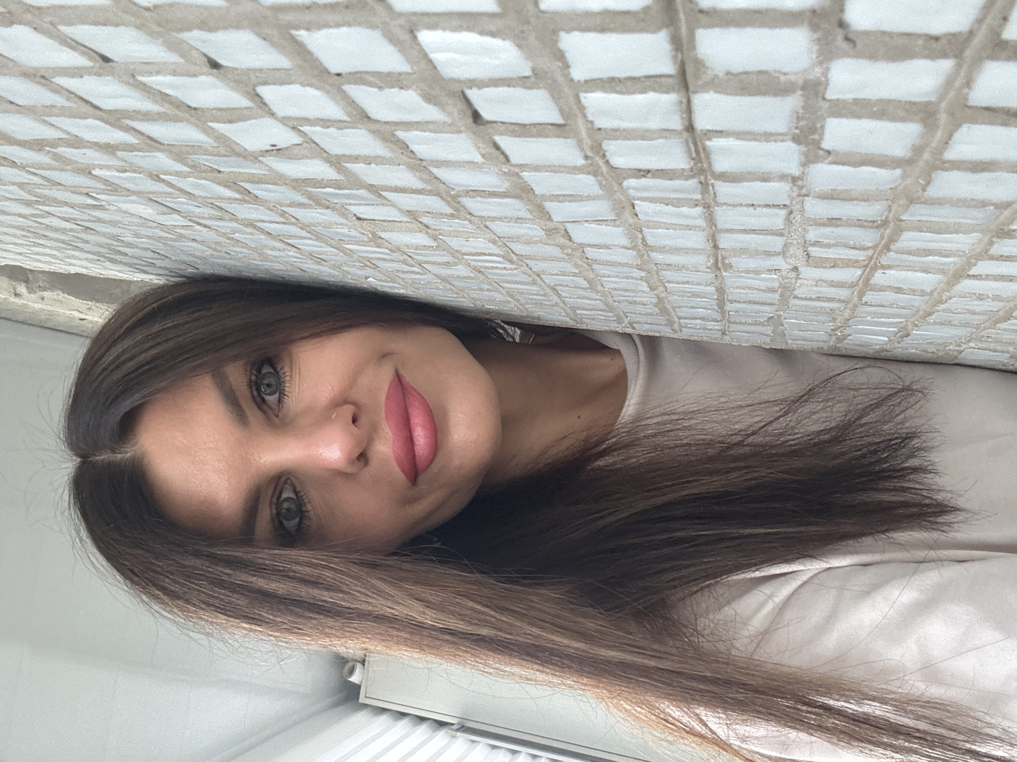 A woman with long, dark brown hair and light-colored eyes is leaning against a tiled wall. She is wearing a light-colored shirt and has pink lipstick on. The tiled wall is white and grey, with a grid pattern. To the left of the woman, a wardrobe or cabinet is visible. The scene appears to be indoors or in a sheltered outdoor area with a bright, natural light source. The location is Chaikovsky, Russia.