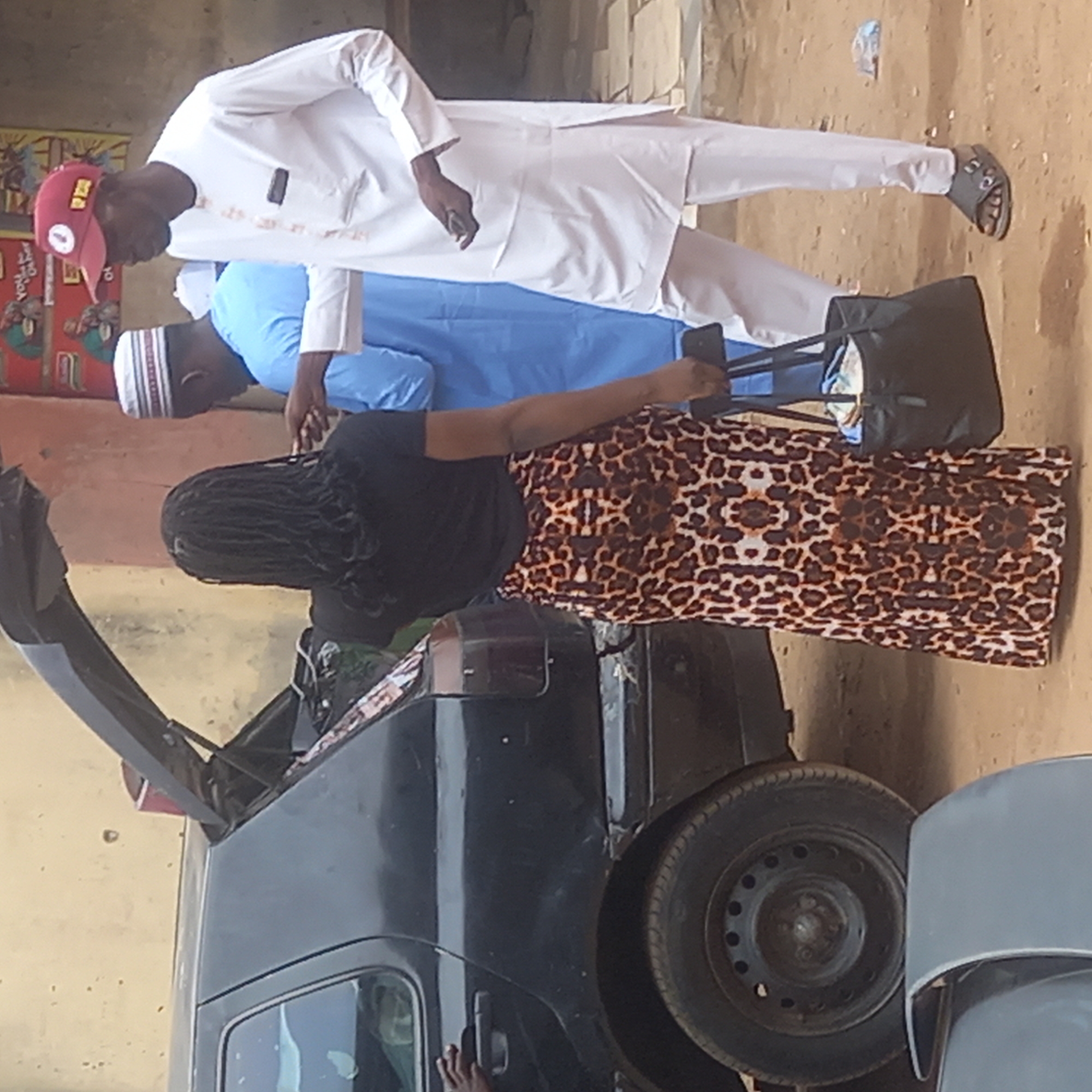 The scene shows three individuals standing near a dark-colored car with its trunk open. The location appears to be outdoors, possibly a roadside in Dadi Riba, Nigeria.
A woman with braided hair is positioned close to the car, wearing a black top and a long, leopard-print skirt, holding a dark-colored mobile phone. Two men stand beside her. One man is dressed in a white outfit and a red cap, while the other is in a light blue outfit and a white cap. A black bag is seen held by the man in the light blue outfit. The ground appears to be a mixture of dirt and possibly a paved surface. A wall is visible in the background with possible commercial advertising.