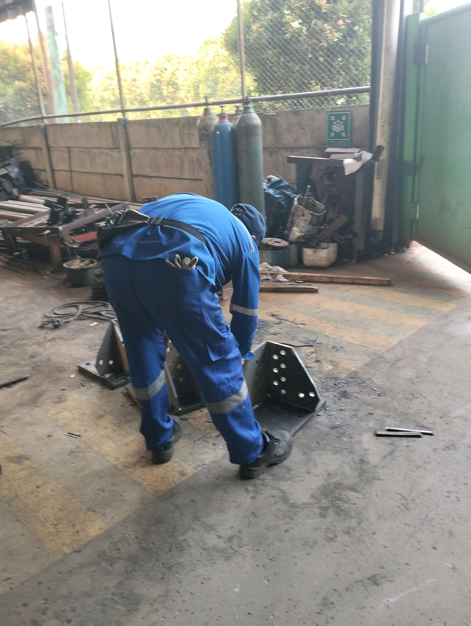 A person wearing blue work overalls with reflective stripes on the legs is bent over in a workshop, appearing to be working on a metal object. The person is wearing black work boots. A tool belt is visible around their waist, with some tools tucked into it. The environment suggests an industrial setting, with concrete flooring, a wall in the background, and various metal objects and equipment scattered around. Two large gas cylinders are visible in the background, one blue and one gray, along with a green metal door. The lighting suggests it may be daytime. The location is described as a workshop in Gunung Sindur, Indonesia.