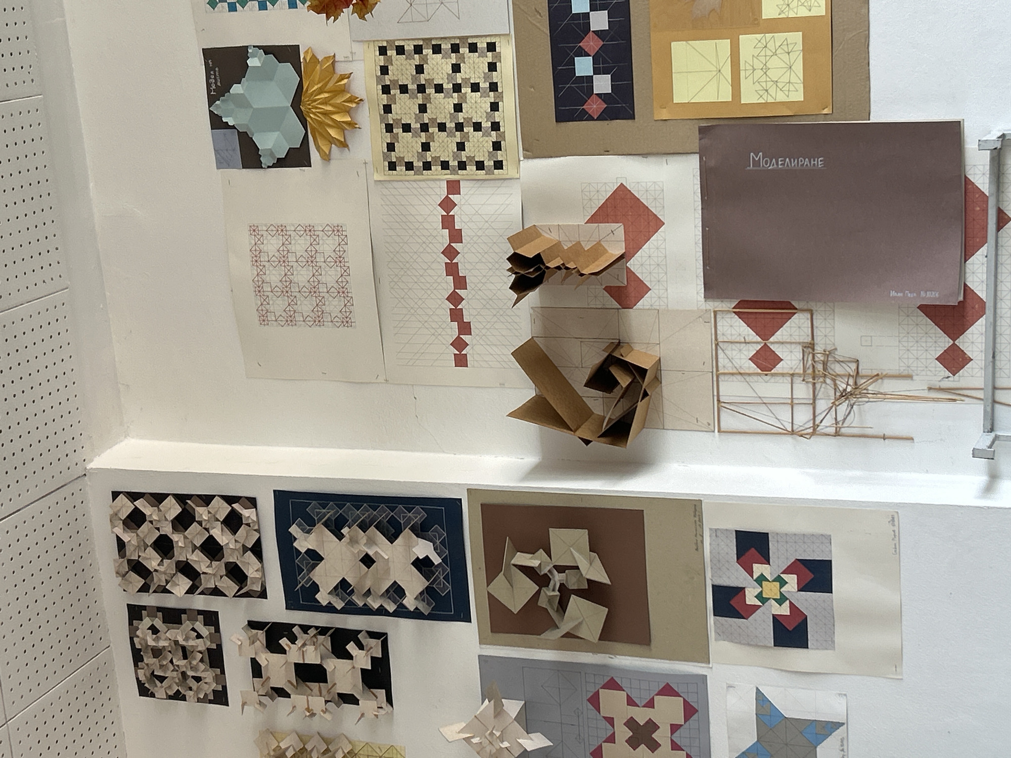 This media file presents a wall display of geometric art and design projects, likely from an educational institution in Sofia, Bulgaria. The collection features a blend of two-dimensional prints and three-dimensional paper models, showcasing studies in pattern, tessellation, and modular construction.
The white wall is adorned with various printed sheets and posters. These include intricate geometric patterns with repeating motifs of squares and diamonds in colors like red, gray, and white. One print displays a checkerboard pattern of dark and light squares. Another shows a multi-faceted, light blue 3D shape against a dark background, with the legible text "Modos en Japonés." Other prints resemble quilt squares, incorporating abstract designs with reds, blues, yellows, and greens.
Among the 2D works are several folded paper sculptures, predominantly in brown cardboard tones, demonstrating complex forms and spatial arrangements. A striking golden, spiky, origami-like model, resembling a sun or flower, stands out. A delicate structure crafted from thin wooden sticks, possibly an architectural or design model, is also part of the display.
A prominent dark brown folder or book is titled "МОДЕЛИРАНЕ" (MODELIRANE), which translates to "MODELING," written in Cyrillic. Below this, smaller Cyrillic text reads "Иван Гоев № Ю20С" (Ivan Goev No. Yu20S), likely identifying the student and project.
A white perforated panel borders the wall on the left. The scene, devoid of people, suggests an exhibition or presentation of student work, focusing on architectural design, fine art, or applied geometry. The overall impression is one of creative exploration and technical skill in visual design.