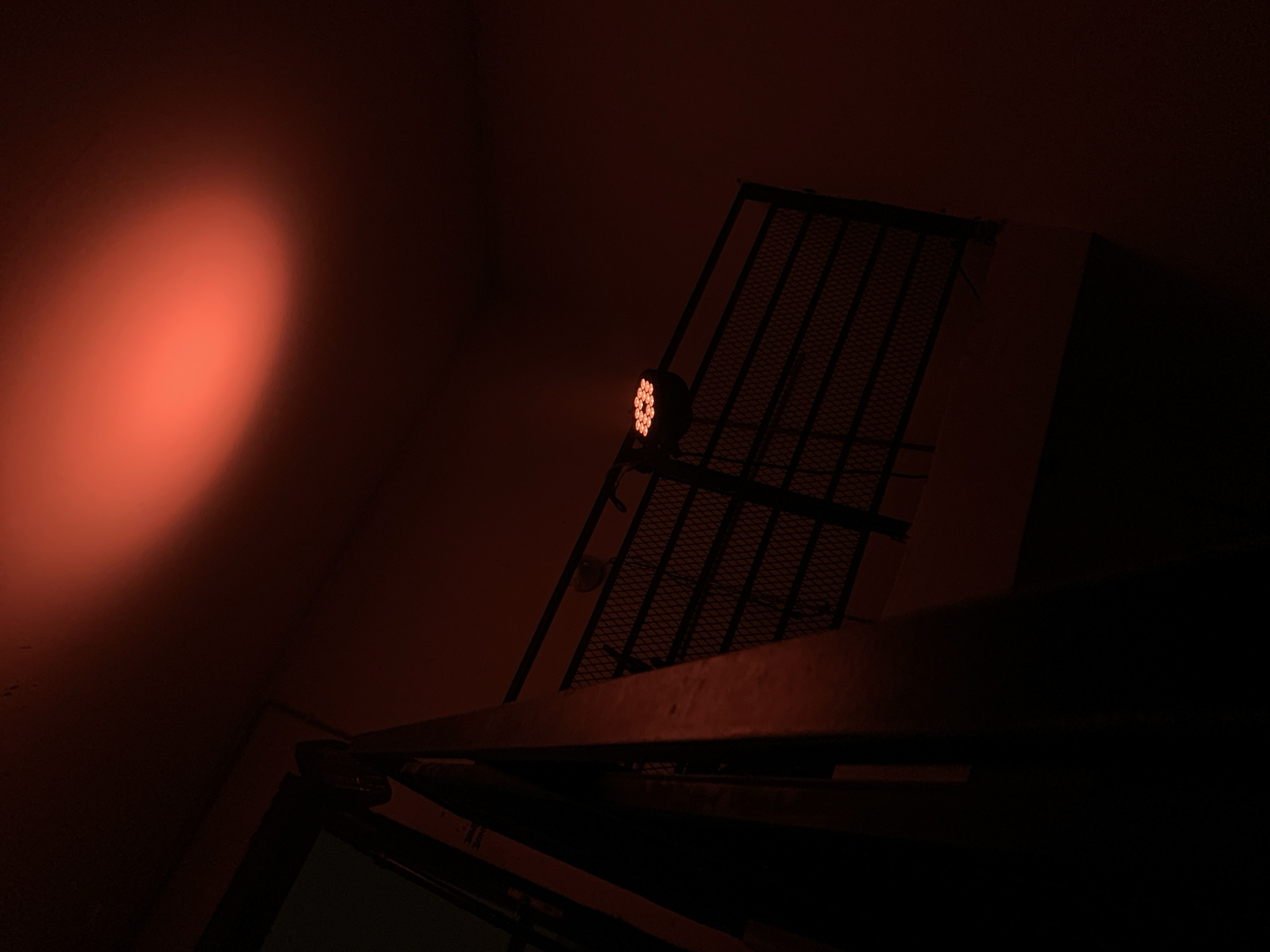 The image depicts a dimly lit interior space, likely in Tel-Aviv, Israel, given the prompt's instruction. A wall with a diffused red light source on the left illuminates the scene. A metal grate structure, possibly part of a fire escape or industrial equipment, occupies the center and right of the frame. An LED light fixture is attached to the grate. The scene is dark, with details obscured by the low light conditions. A portion of an open structure is in the foreground.
