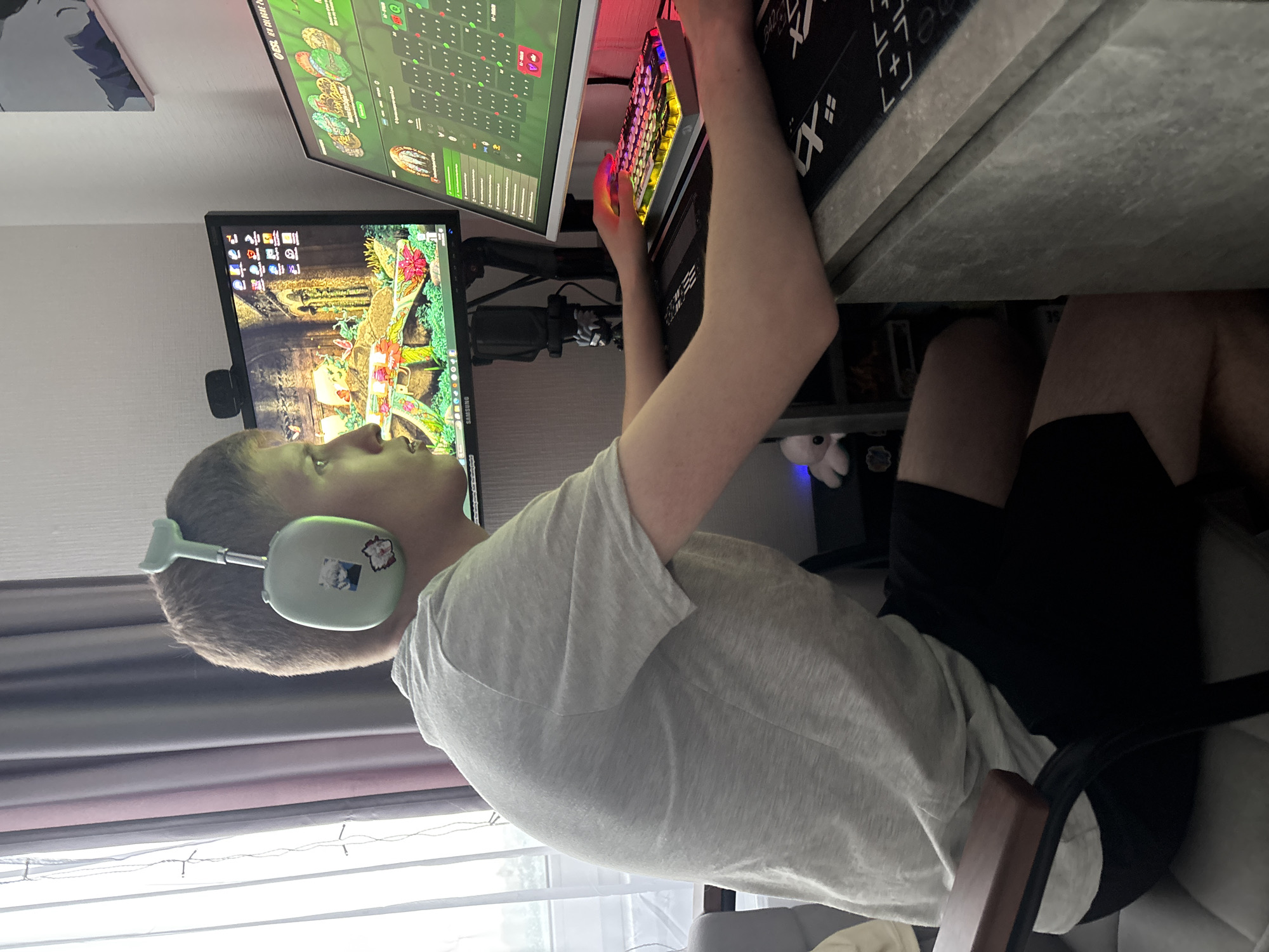 A young male sits at a desk in front of two computer monitors, wearing a light-colored T-shirt, dark shorts, and light green headphones adorned with stickers. He is seated in a chair with a wooden armrest. His hands are positioned on a colorful keyboard. The monitors display digital images, one appearing to show a map or strategic interface, and the other showing what looks to be a desktop with icons and a nature-themed background. A microphone is positioned near the monitors. A desk with a patterned mousepad is visible. The location is inferred to be in Vsevolozhsk, Russia.