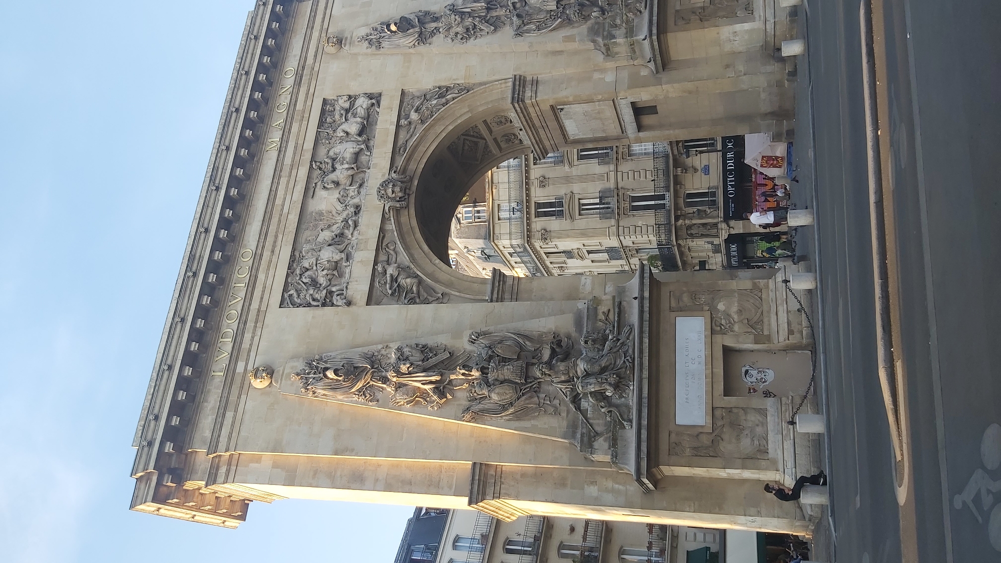 The image shows the Arc de Triomphe du Carrousel in Paris, France, viewed from the side. The arch is made of stone and features intricate sculptures and inscriptions. The inscription "LVDV.ICO MAGNO" is visible at the top of the arch. Below the inscription, a relief depicts a battle scene with figures on horseback. On the left side of the arch, there is a large sculptural panel with several figures and armor.
In the background, through the archway, several multi-story buildings with typical Parisian architecture are visible. Some of these buildings have balconies and ornate facades. The sky is clear and blue, indicating a daytime scene.
In the foreground, a street with a road and sidewalk is present. A person is sitting on a bollard on the sidewalk to the left of the arch. Several other people are visible in the background on the sidewalk, some standing and some walking. There are also some shops with signage, including "OPTIC DUROC." The overall lighting suggests it is late afternoon or early evening.