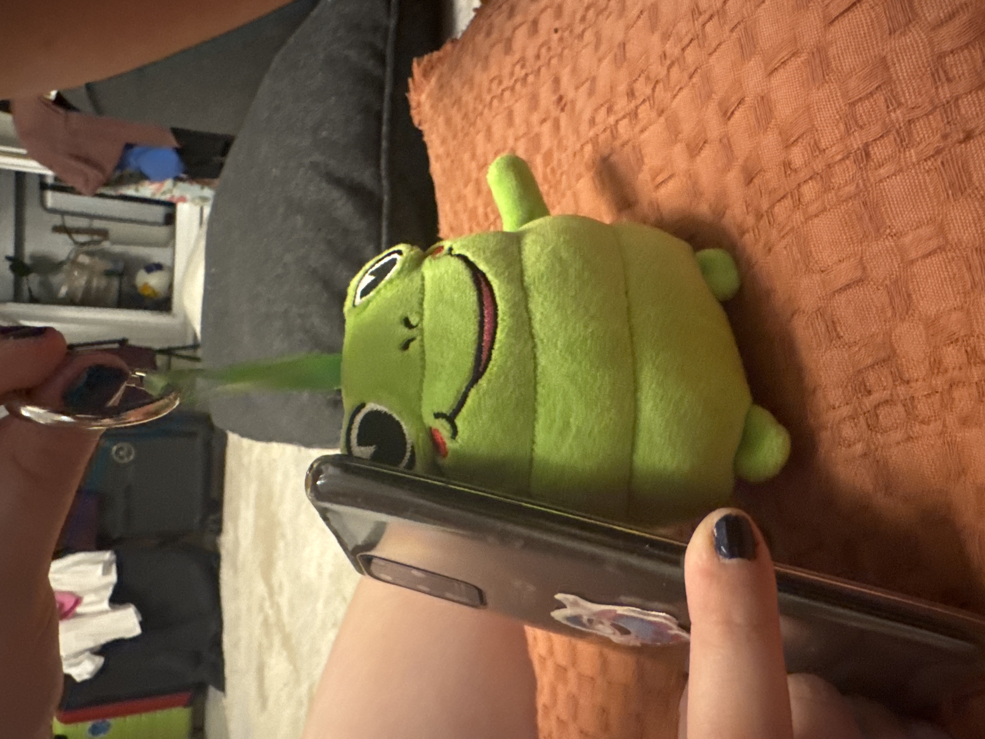 A close-up, slightly overhead shot shows a hand holding a phone, with a plush frog toy leaning against the phone. The frog is bright green with large black eyes, rosy cheeks, and a wide smile. It is leaning on an orange textured surface. In the background, a dark gray pillow is visible, as well as what appears to be luggage and clothing. The scene is indoors, with the lighting suggesting it may be evening or a dimly lit room. The image is cropped in a way that focuses on the phone and the toy, obscuring the full context of the environment.