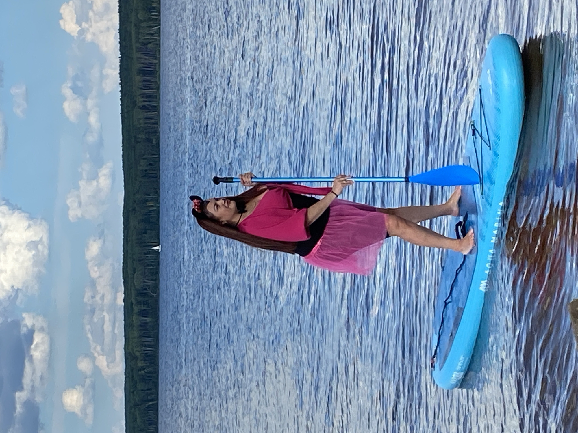 The image shows a person on a stand-up paddleboard in a body of water. The person is wearing a pink top and a pink tutu. They are holding a blue paddle and are looking upwards. A blue paddleboard with black bungee cords is visible. In the background, there is a tree line and a blue sky with white clouds. A small sailboat is visible in the distance. The water appears calm with ripples. The scene is illuminated by natural light, suggesting it is daytime. There is text visible on the paddleboard which reads "AQUA MARINA". The image is oriented vertically. The location is Chaikovsky, Russia.