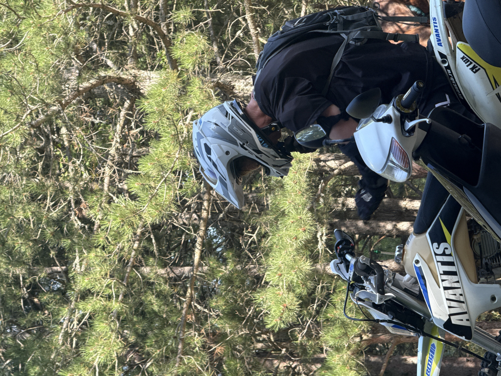 An outdoor scene captured from a slightly low angle features a person wearing a white and grey motocross helmet and a black t-shirt, sitting on a dirt bike. The person is looking down towards the handlebars of the bike. A black backpack is visible on their back. The dirt bike itself is predominantly white and black with blue and yellow accents, and the brand name "AVANTIS" is prominently displayed on its side. The background is dominated by a dense forest of pine trees, with sunlight filtering through the branches, creating a dappled lighting effect. The ground is not clearly visible, but appears to be a natural, unpaved surface, consistent with a forest environment. The overall atmosphere is casual and recreational.