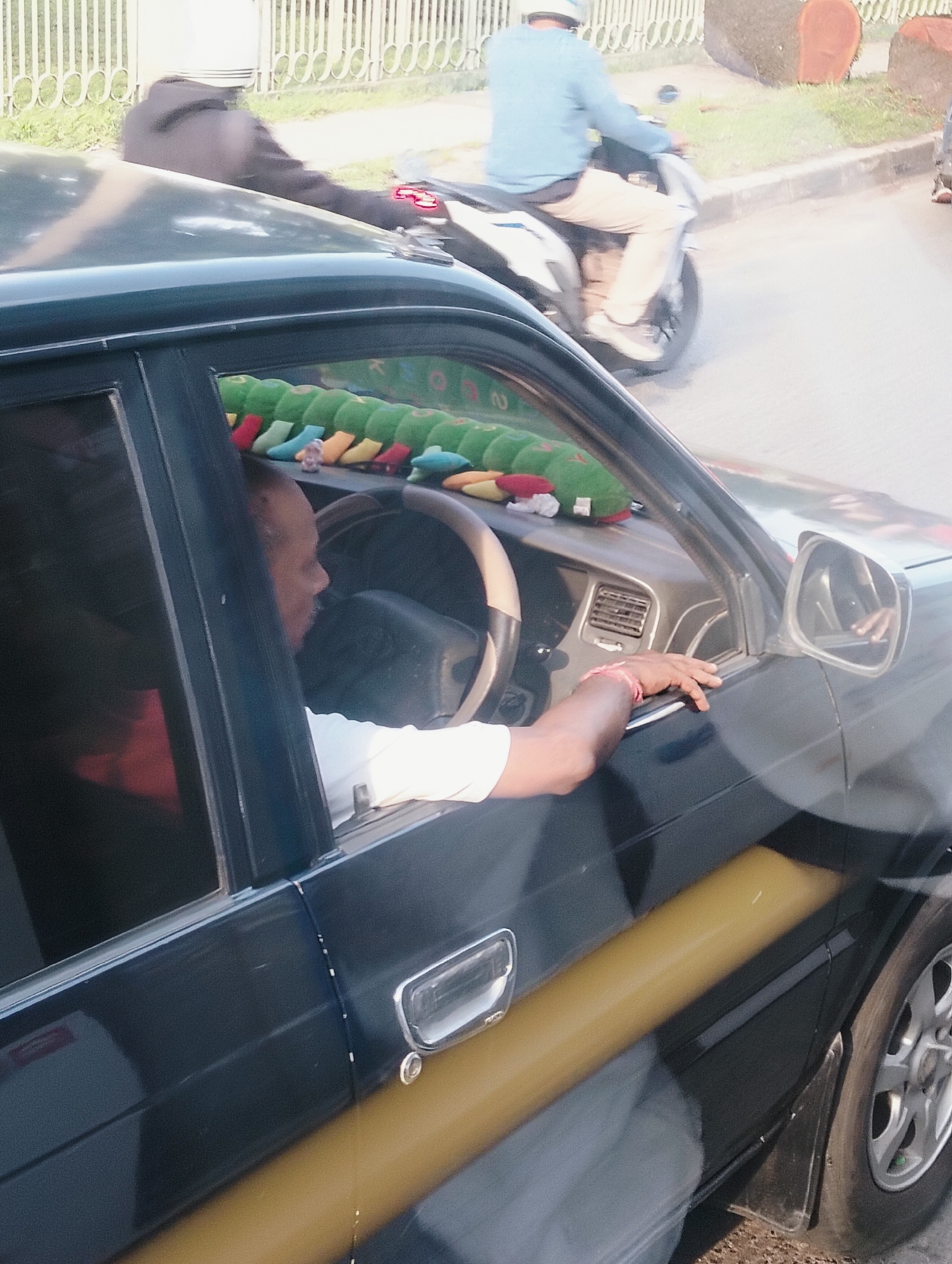 In the City of Medan, Indonesia, a dark-colored car is parked, with its driver's side door open. A man with dark skin is visible inside the car, leaning his left arm on the doorframe and his right hand on the steering wheel. He is wearing a white t-shirt. The car's interior features a steering wheel with a tan-colored rim and a dashboard with an air vent. A string of brightly colored, caterpillar-shaped plush toys is draped across the dashboard. Outside the car, a person wearing a blue long-sleeved shirt and light-colored pants is riding a scooter, their face obscured by a helmet. In the background, a white metal fence runs along a grassy area, and two large, round logs are visible. The scene appears to be on a street with a paved surface.