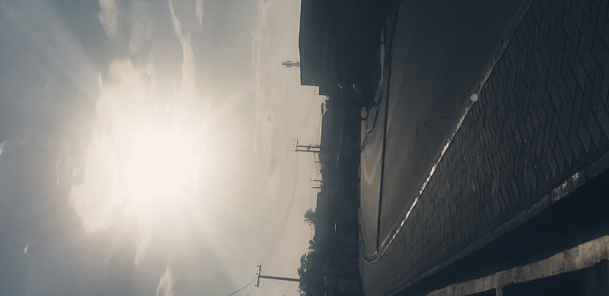 The image depicts a street scene in Narayi, Nigeria, characterized by a bright, overcast sky with the sun partially obscured by clouds, casting diffuse light. In the foreground, a paved road curves gently to the right, bordered by a brick-patterned walkway. Overhead, power lines are visible, supported by utility poles. Silhouetted buildings and structures form the backdrop, with a prominent cell tower on the right side of the frame. The scene appears to be a quiet, possibly early morning or late afternoon moment, with no people or vehicles readily apparent. The overall impression is one of an urban street in a tropical climate, captured under atmospheric lighting conditions.