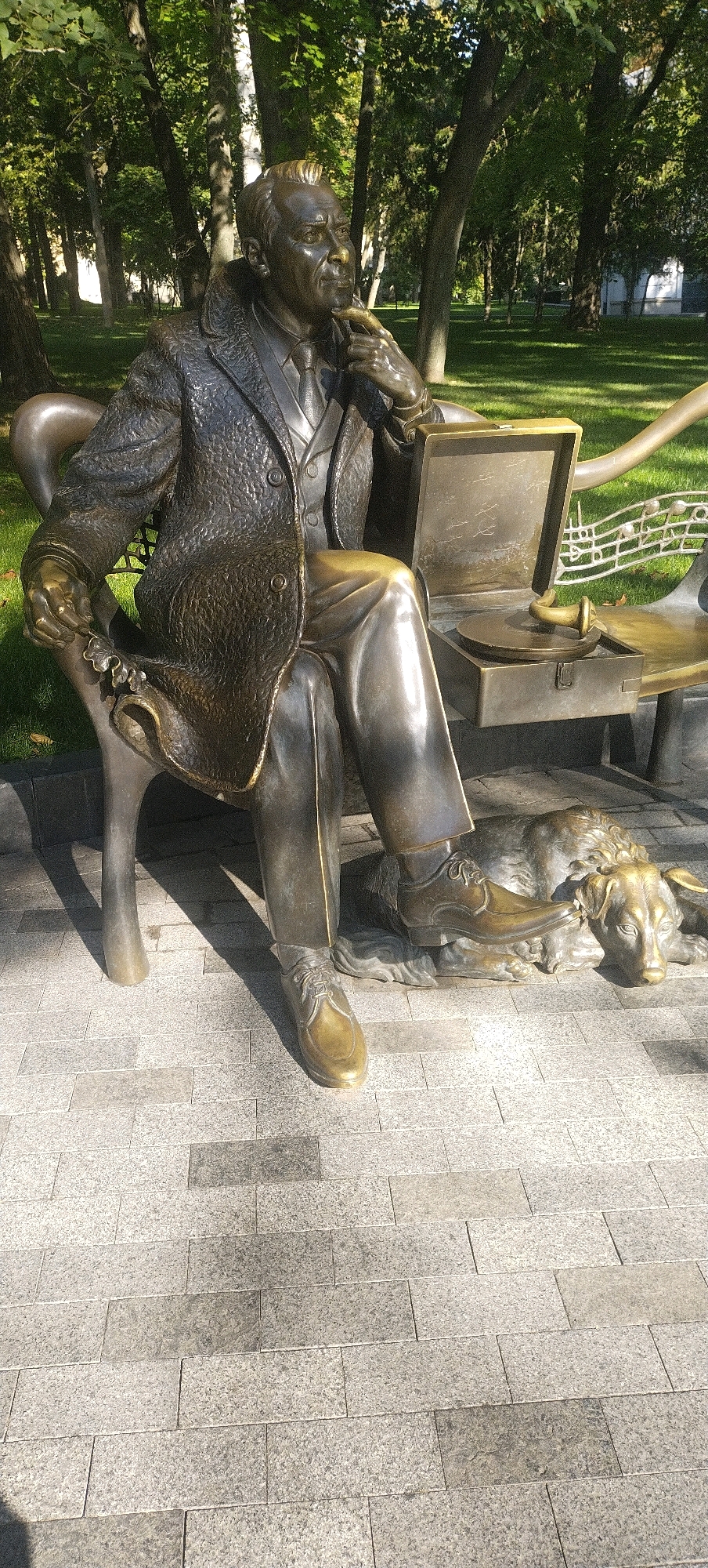 A bronze sculpture of a man sitting on a bench in a park depicts him wearing a suit, tie, and trench coat, with his right hand near his chin and his left hand holding a small object. Next to him on the bench is an open portable record player. A bronze dog is lying on the ground at his feet. The sculpture is located in Kharkiv, Ukraine. The background shows trees and grass, suggesting a park setting on a sunny day. The ground in the foreground is paved with rectangular stones.
