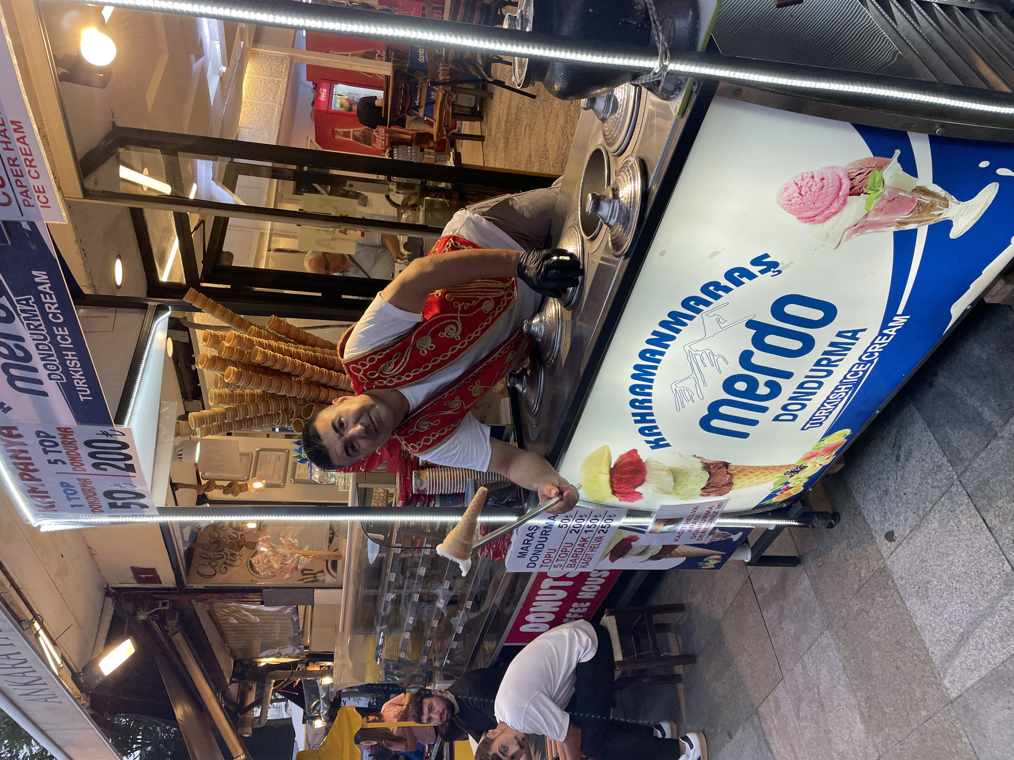 In Kadıköy, Turkey, a vendor wearing a red vest over a white shirt and black gloves presents a cone of Turkish ice cream. The vendor stands behind a refrigerated display of ice cream, which has a large blue and white graphic featuring the words "KAHRAMANMARAS" and "merdo DONDURMA TURKISH ICE CREAM." Several other cones of ice cream are stacked on the counter. Two men are seated to the left of the vendor, one in a white t-shirt and the other in a black t-shirt. In the background, a Coca-Cola refrigerator is visible. The scene appears to be outdoors, with a cobblestone-like ground surface.