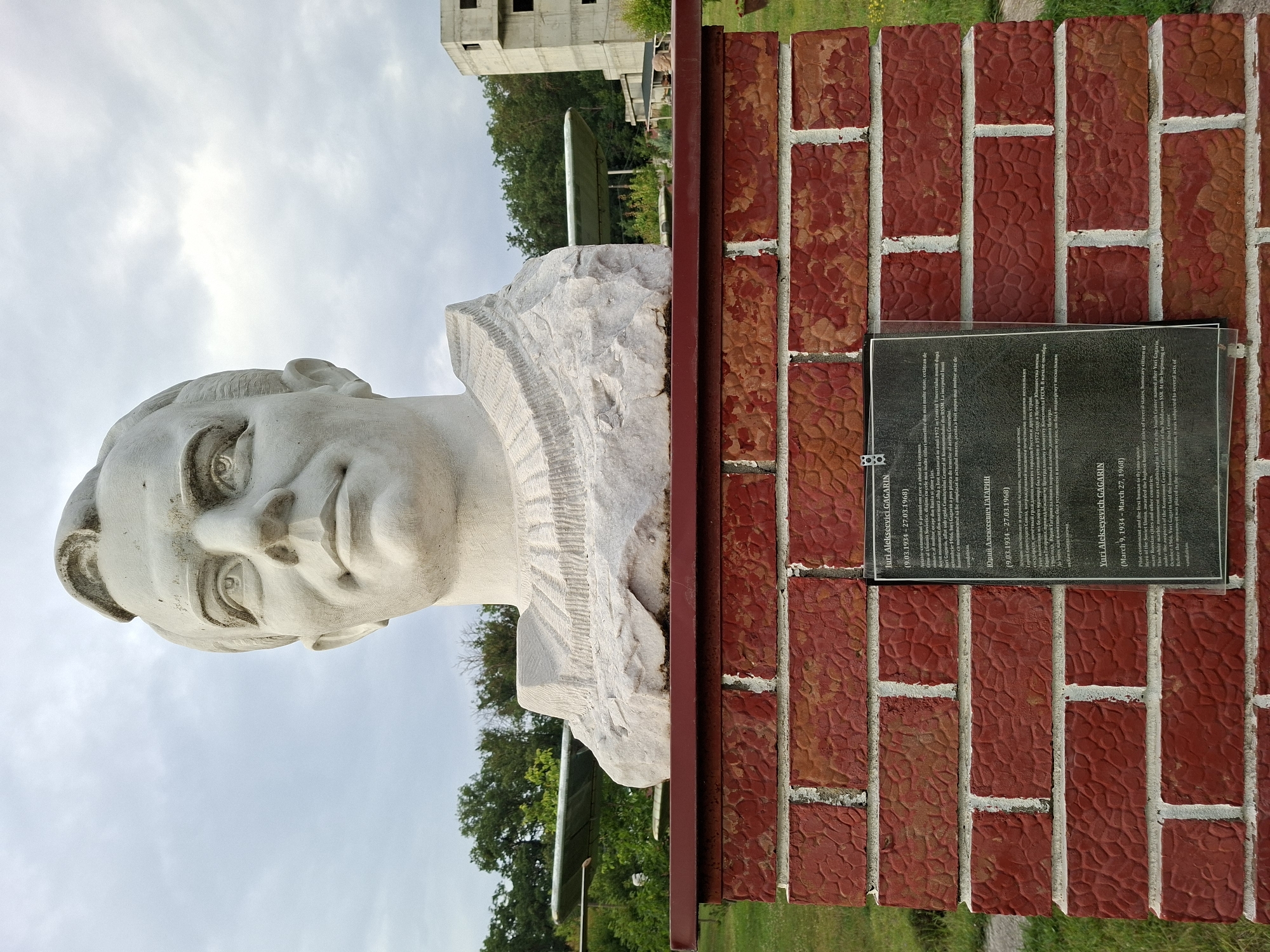 This image depicts a bust of Yuri Gagarin, the first human in space, on a brick pedestal. The bust is made of white stone and is mounted on a red brick structure. A plaque with biographical information about Gagarin is affixed to the brick pedestal, presented in Romanian, Russian, and English. Behind the bust, a partially constructed building with multiple floors and exposed concrete is visible, along with trees and a cloudy sky. The presence of what appears to be an airplane wing or similar structure in the background suggests the location might be near an airfield or aviation-related site. The general setting appears to be an outdoor park or memorial area. The scene is captured from a slightly low angle, looking up at the bust. The city of Malcoci, Moldova, is the location of this monument, specifically the House of Culture, which houses a museum dedicated to Gagarin.