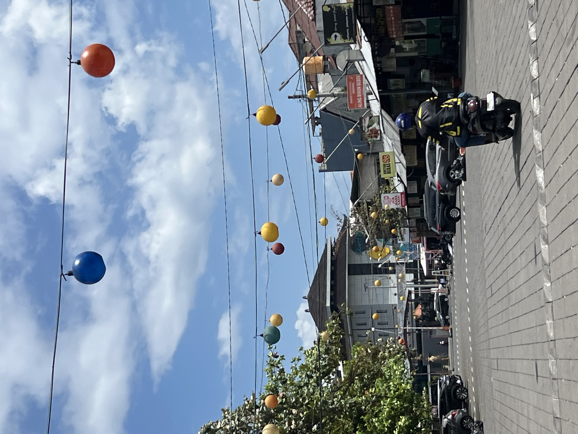 The image depicts a street scene in Kuta, Indonesia, characterized by a bright, sunny day with scattered clouds. Overhead, a series of colorful spherical lanterns are suspended from wires, including prominent blue and orange ones against the sky, and a scattering of yellow, green, and red lanterns further down the street. The street itself is paved with grey brickwork, marked with white lines.
Several vehicles are present, including a silver car parked on the left, a dark SUV, and a dark sedan in motion. A person wearing a black jacket with yellow accents and a blue helmet is riding a scooter away from the camera, positioned in the center of the street.
The background features buildings with dark tiled roofs, some appearing to be shops or restaurants, with signage visible. One sign reads "ALING BIKUN BEBEK." A lush green tree with dense foliage is on the left side of the image, partially obscuring some of the buildings. The overall impression is a lively street, likely a commercial area in a tropical setting, decorated for an event or as part of the local ambiance.