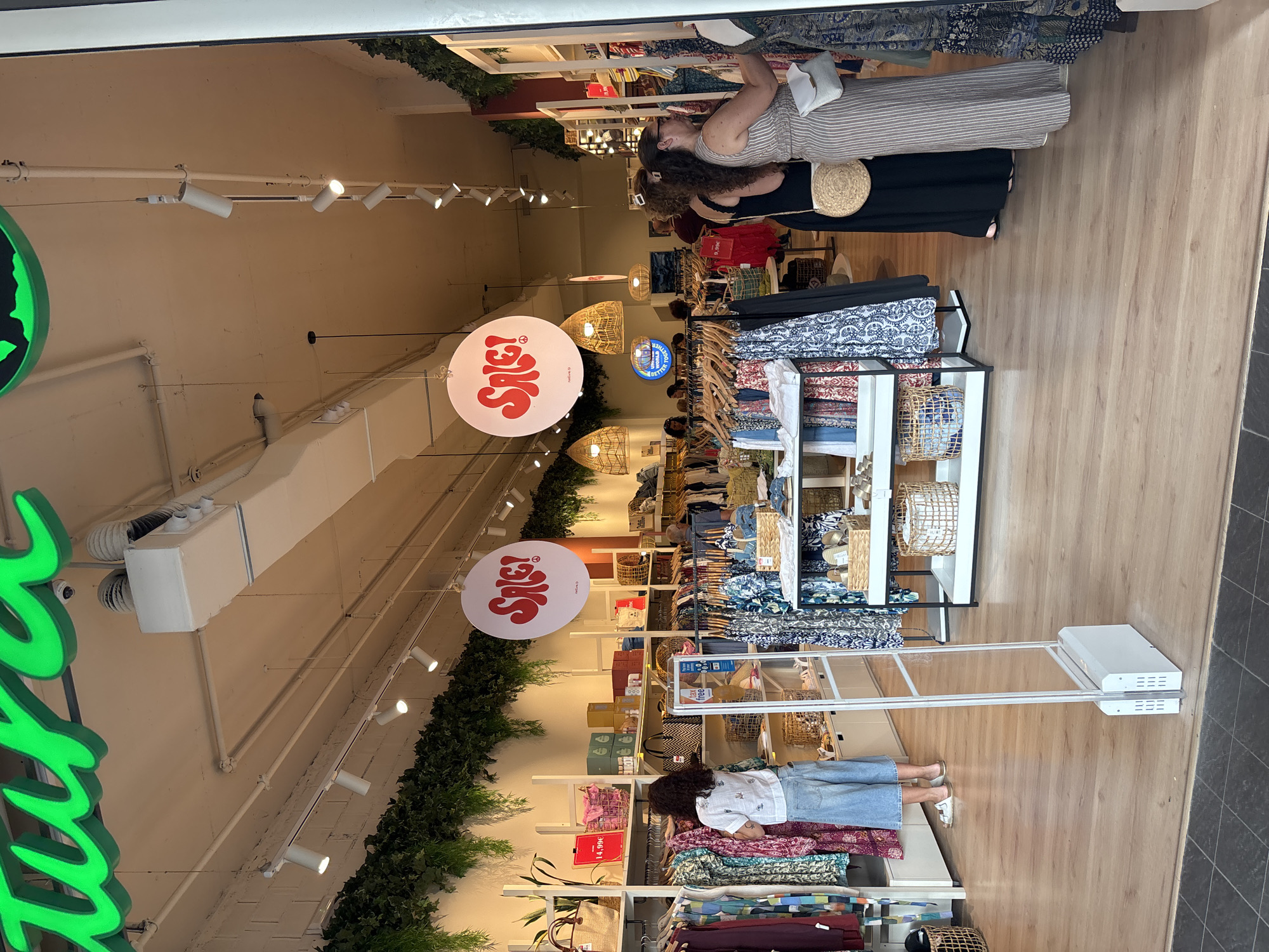 The image shows the interior of a clothing store, likely located in Orihuela, Spain, given the "Natur" signage which is a prominent brand in the region. The store features a bright and airy atmosphere, with clothing racks filled with apparel, display shelves with accessories and decorative items, and hanging light fixtures. The flooring is a light-colored wood.
In the foreground, a security gate is visible, indicating an entrance or exit point. To the left, a woman with curly dark hair wearing a white top and wide-leg denim pants is browsing a rack of colorful dresses. She is facing away from the camera.
Further into the store, several other individuals are present. Two women are seen in the middle ground, one in a black dress and the other in a striped maxi dress, both appearing to examine clothing. In the background, more shoppers are browsing the merchandise. Large, circular signs with the word "SALE!" in red are hanging from the ceiling, announcing a promotion. Green artificial foliage is draped along the top of the walls and ceiling, adding a touch of natural decor. The overall scene depicts a typical retail environment focused on women's fashion.