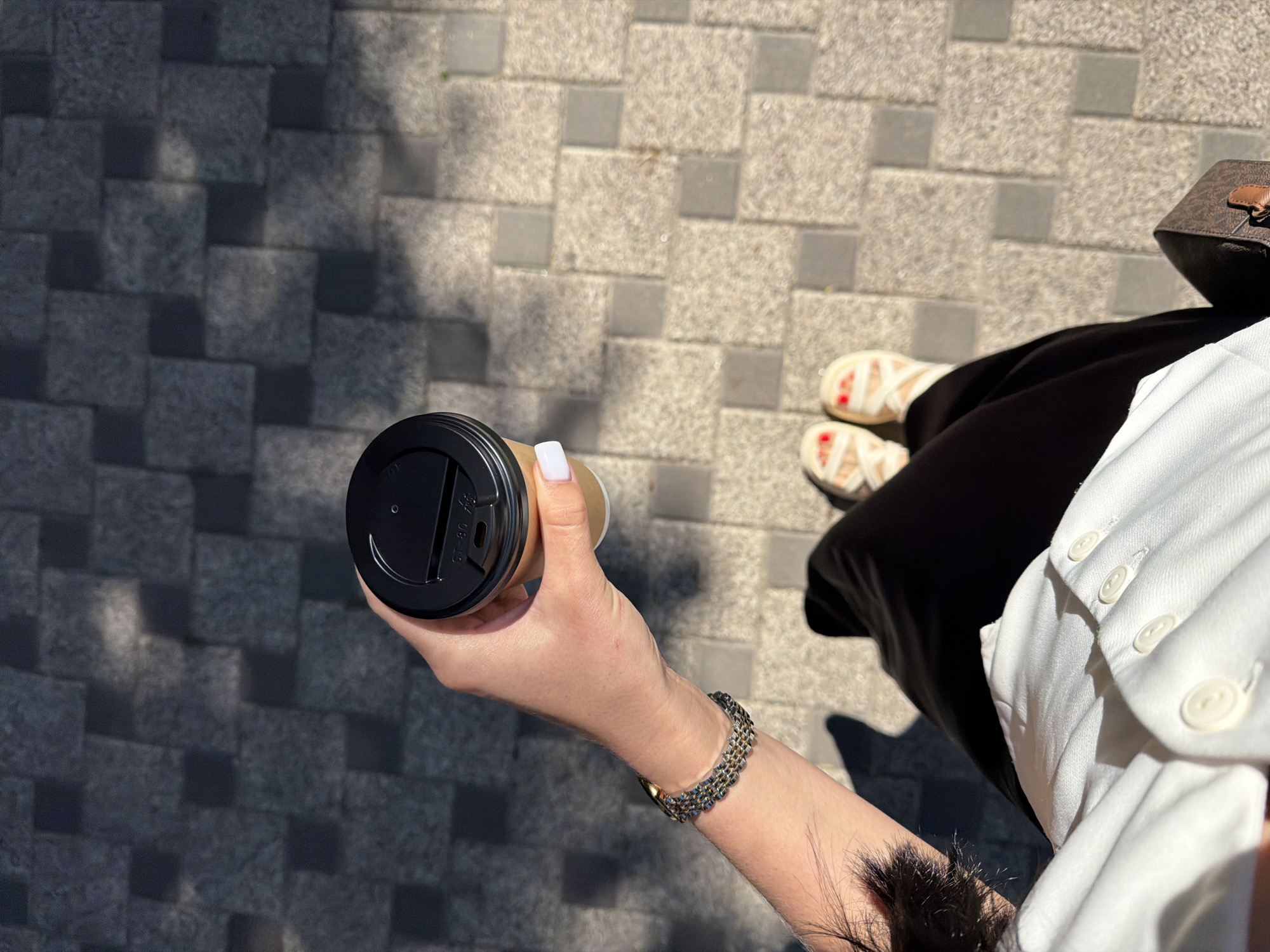A person, seen from a high angle, holds a disposable coffee cup with a black lid. The person's right hand, with neatly manicured white nails, is wrapped around the cup. A silver and gold-toned bracelet adorns their wrist. They are wearing a white collared shirt and black pants. White sandals with crisscross straps are visible on their feet, and red nail polish is apparent on their toes. The scene takes place outdoors on a paved surface composed of grey and tan square tiles, interspersed with darker grey tiles, arranged in a grid pattern. Sunlight casts shadows across the pavement. A dark brown handbag with a gold buckle rests near the person's right leg. The overall impression is of a person casually walking outdoors on a sunny day.