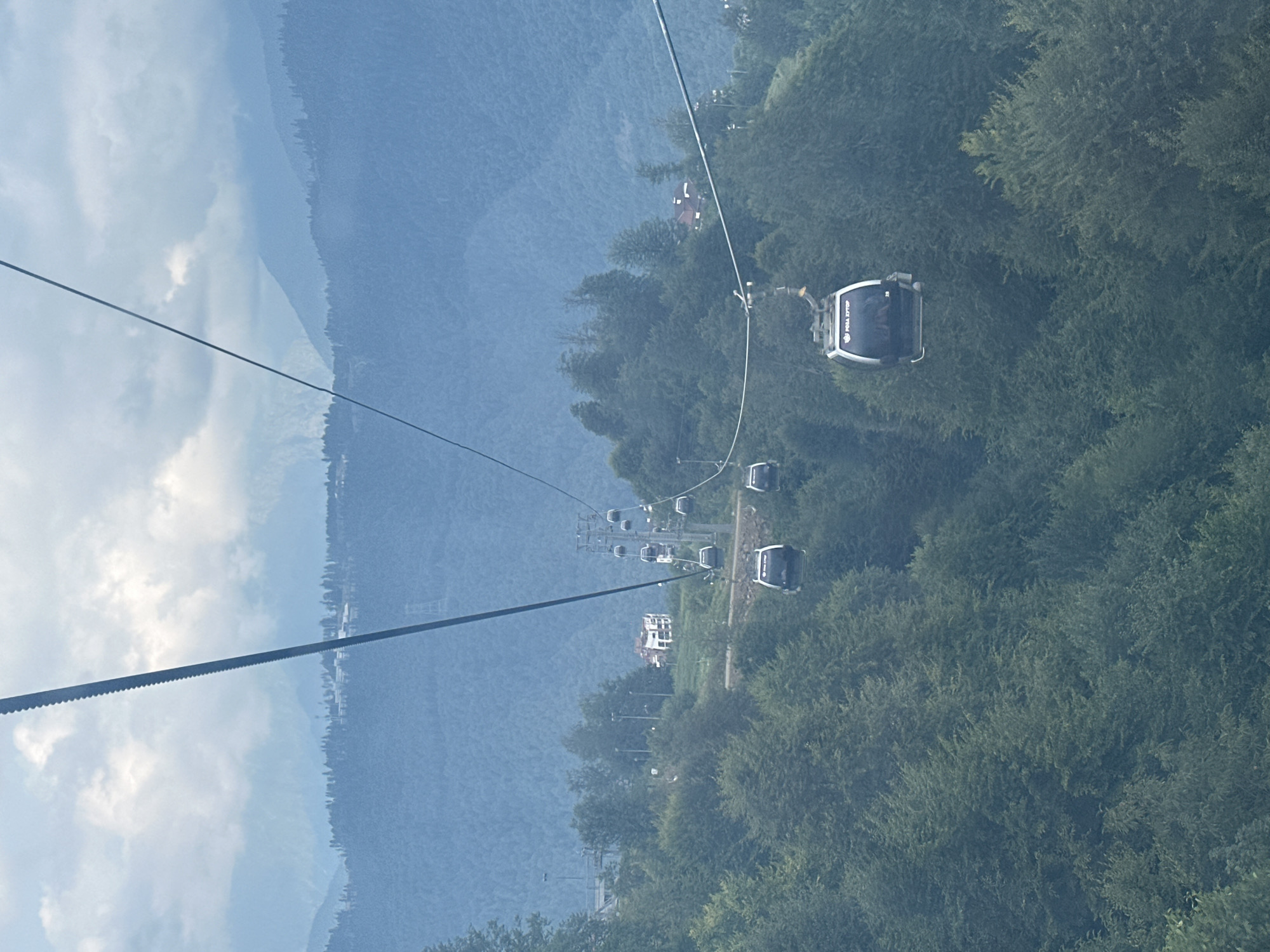 This image depicts a mountainous landscape with a cable car system, likely in Estosadok, Russia, a resort town known for its ski slopes and scenic views. The cable car, with multiple cabins suspended on a wire, ascends a steep, densely forested slope. The cabins appear to be modern, with some featuring illuminated signage. The surrounding environment is characterized by lush green trees covering the mountainside, with a few buildings visible in the distance, possibly part of a village or resort infrastructure. The sky is overcast with clouds, casting a soft light over the scene, and further mountain ranges are visible in the background, fading into a hazy blue. The perspective is from a higher vantage point, looking down and along the cable car line.
