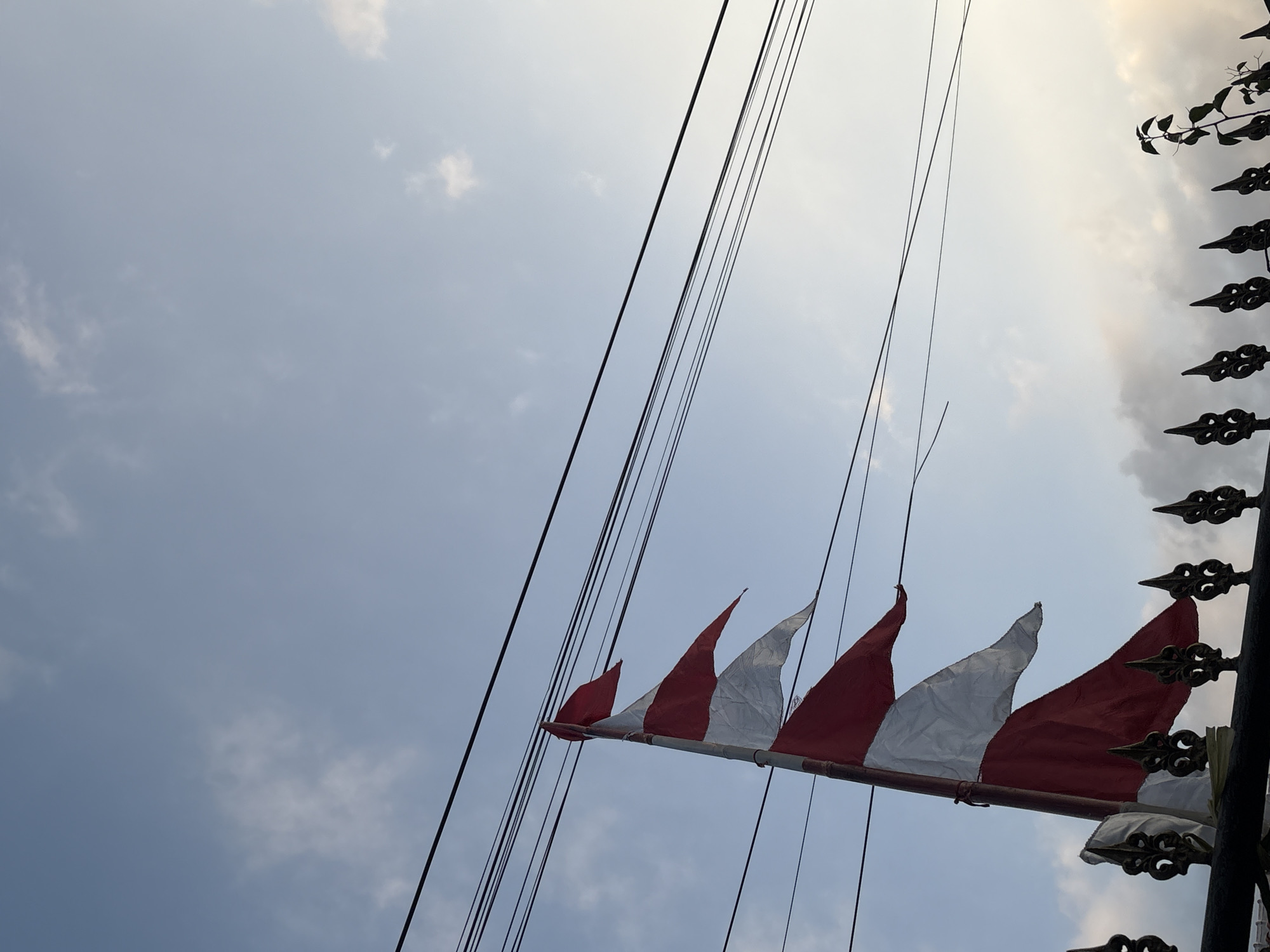 A low-angle, full-shot photograph captures a string of red and white triangular flags attached to a pole. The flags are fluttering in the wind against a pale blue sky dotted with wispy white clouds. Multiple black power lines are strung horizontally across the frame above the flags. In the foreground, a decorative black metal fence is visible, with a small green plant growing on its right side. The image appears to be taken in Palimanan, Indonesia.