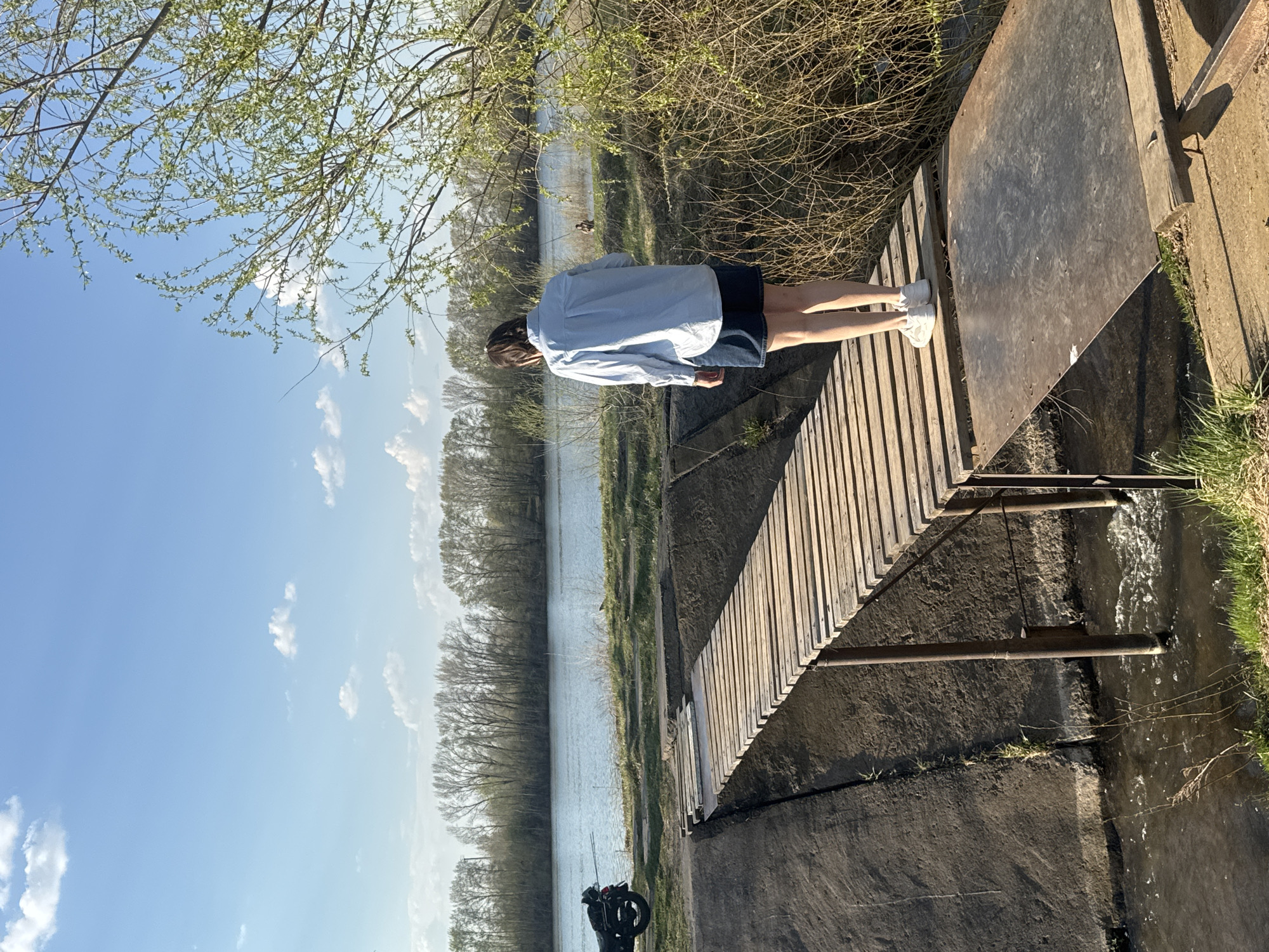 A person stands on a wooden bridge extending over a canal in Chaikovsky, Russia. The canal runs parallel to a river bordered by trees. The sky is partly cloudy. The person wears a light-colored shirt and dark shorts with white shoes. The bridge has a sheet of metal on one side. A motorcycle is parked on the ground nearby. Water flows through the canal and under the bridge.