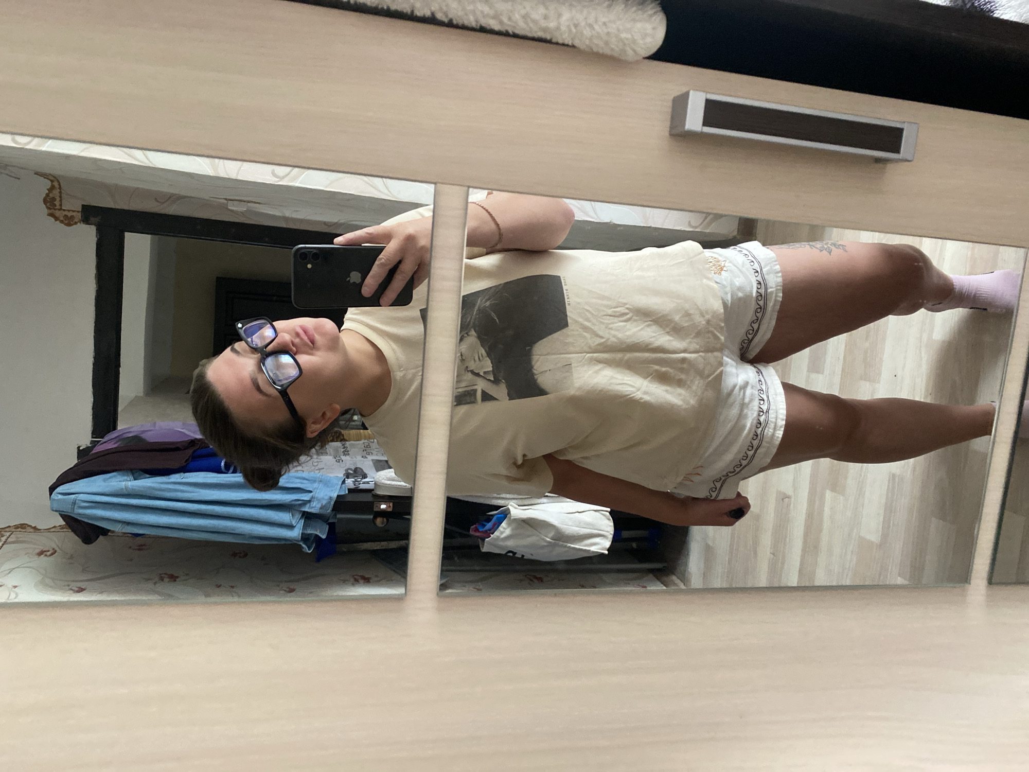 A young woman stands in front of a mirror, taking a selfie with her phone. She is wearing glasses, a light-colored t-shirt with a black and white graphic, and white shorts with a decorative trim. She has a tattoo on her left thigh and is wearing pink socks. The room appears to be a bedroom, with clothes hanging on a rack in the background and a closet door to her right. The flooring is light-colored laminate. The image is likely taken in Otchor, Russia.