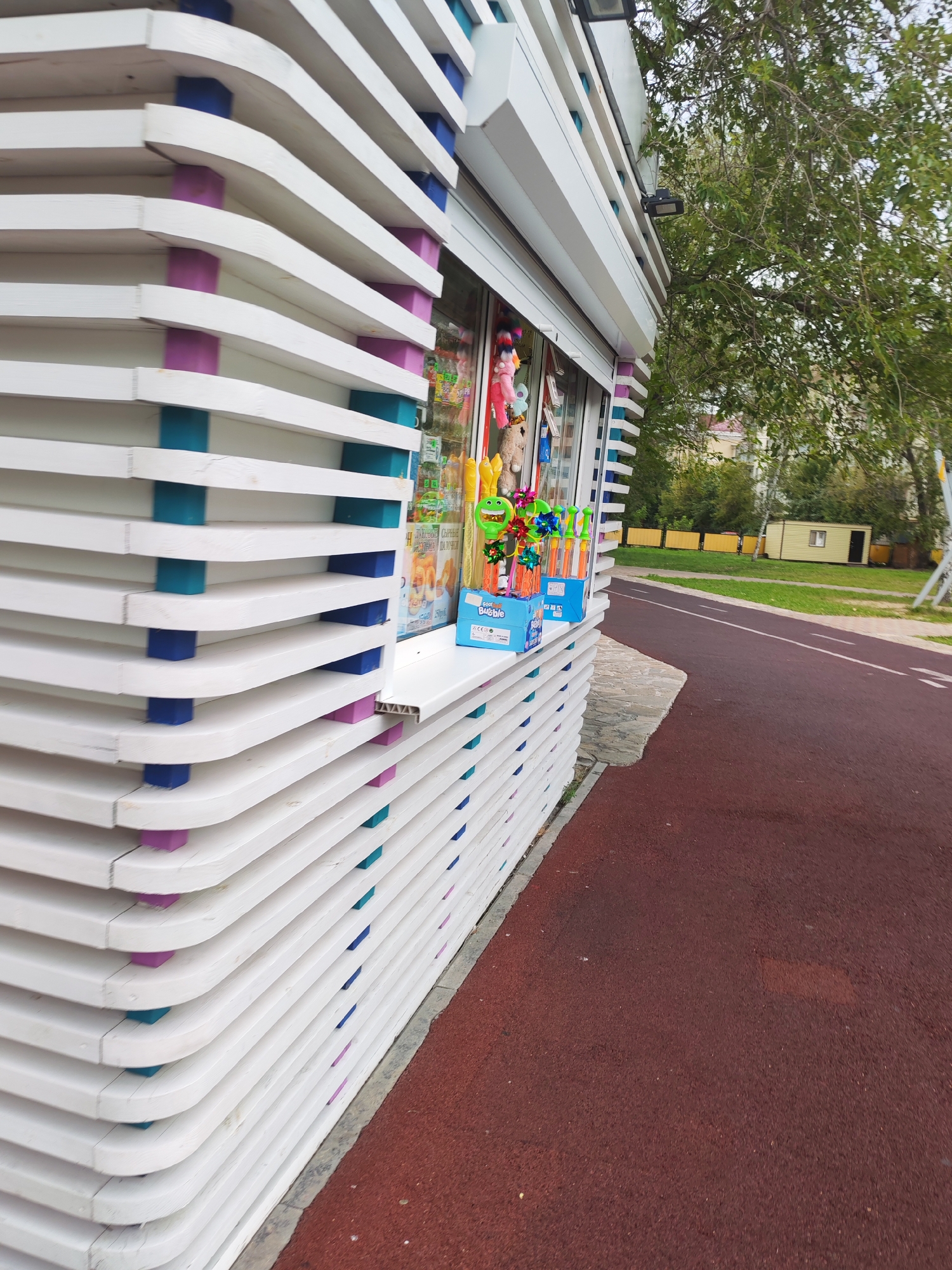 A low-angle, full shot captures a brightly colored outdoor kiosk with white slatted siding, accented with alternating bands of purple and teal supports. The kiosk's facade has a slanted roof and a glass window displaying various toys and candies, including plush animals and bubble wands in blue boxes. To the right of the kiosk is a dark red, asphalt path that curves away into the background, bordered by a grassy area with trees and a small, light yellow building. The sky above is overcast and partially obscured by the green leaves of a large tree growing from behind the kiosk.