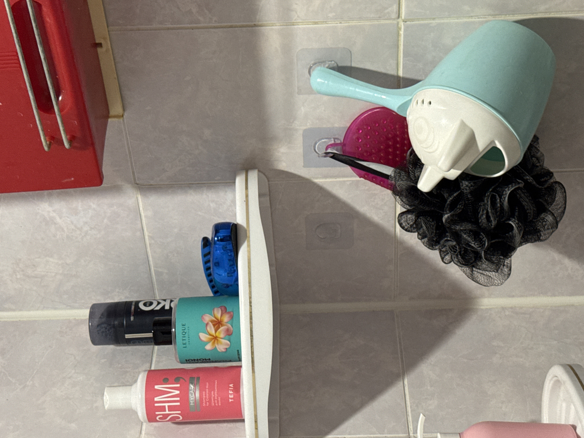 This is a medium-shot photograph, taken indoors, likely in a bathroom. The scene is composed of a tiled wall with a corner to the left. On the left side of the frame, a white shelf with a gold trim holds several items. From left to right, there is a pink bottle with white text that reads "SHM MYCARE Shampoo for Colored Hair," a teal bottle with a flower design and the brand "LETIQUE COSMETICS," and a blue hairbrush. Behind the teal bottle, a tall black aerosol can with white text is visible.
To the right of the shelf, hanging on clear adhesive hooks on the tiled wall, are several bath accessories. A pink textured brush is hanging, and next to it, a black mesh bath pouf is partially visible. A teal and white watering cup, designed for children, is hanging to the right of the bath pouf. Above the shelf, a red plastic rack with two metal bars is mounted on the wall.
The overall environment appears clean and organized. There is no indication of people or any specific actions being performed. The lighting suggests artificial indoor lighting. The location is specified as Яромінскі сельскі Савет, Belarus.