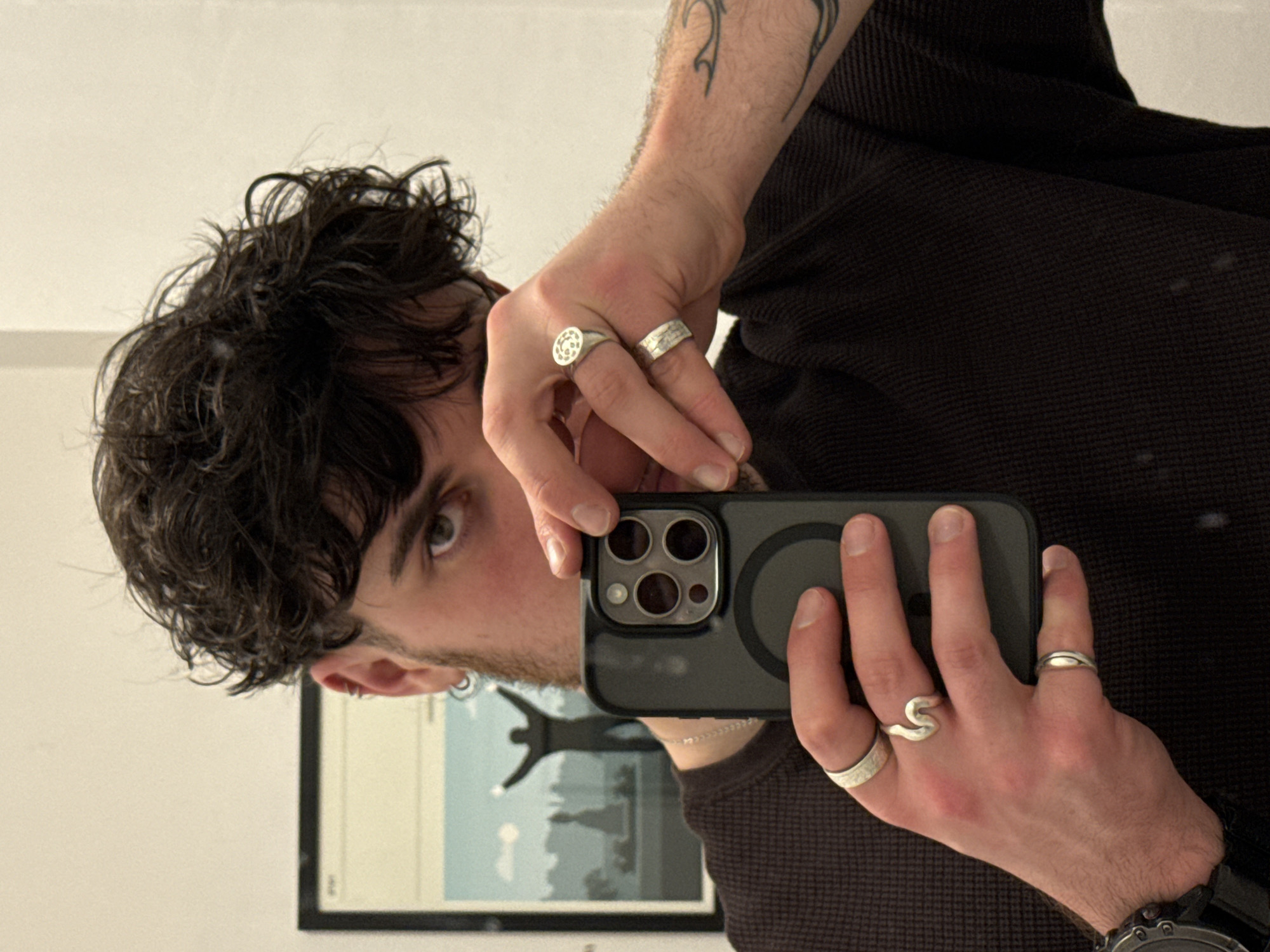 A man takes a selfie in front of a mirror. He has dark, curly hair and is wearing a dark-colored waffle-knit shirt. He is holding a black smartphone with three camera lenses. The man is adorned with multiple silver rings on both hands and a silver earring in his left ear. A tattoo is visible on his left forearm, and a watch on his left wrist. In the background, a framed picture is hanging on the wall. The location is unknown, in an unknown city.