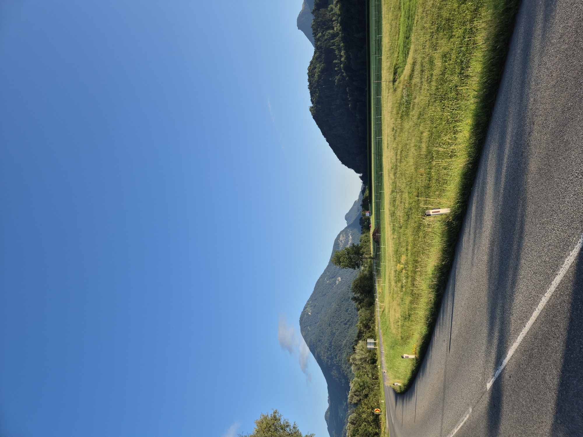 The image shows a scenic view of a curved asphalt road in Niederndorf, Austria. The road has a white line marking the edge and a broken white line in the middle. On the right side of the road, there is a vibrant green grassy field with a white fence. In the background, there are rolling hills covered with dense green trees, and in the distance, majestic mountains rise under a clear blue sky. The sunlight casts shadows on the road, indicating it is likely morning or late afternoon. The overall atmosphere is peaceful and serene, highlighting the natural beauty of the Austrian landscape.