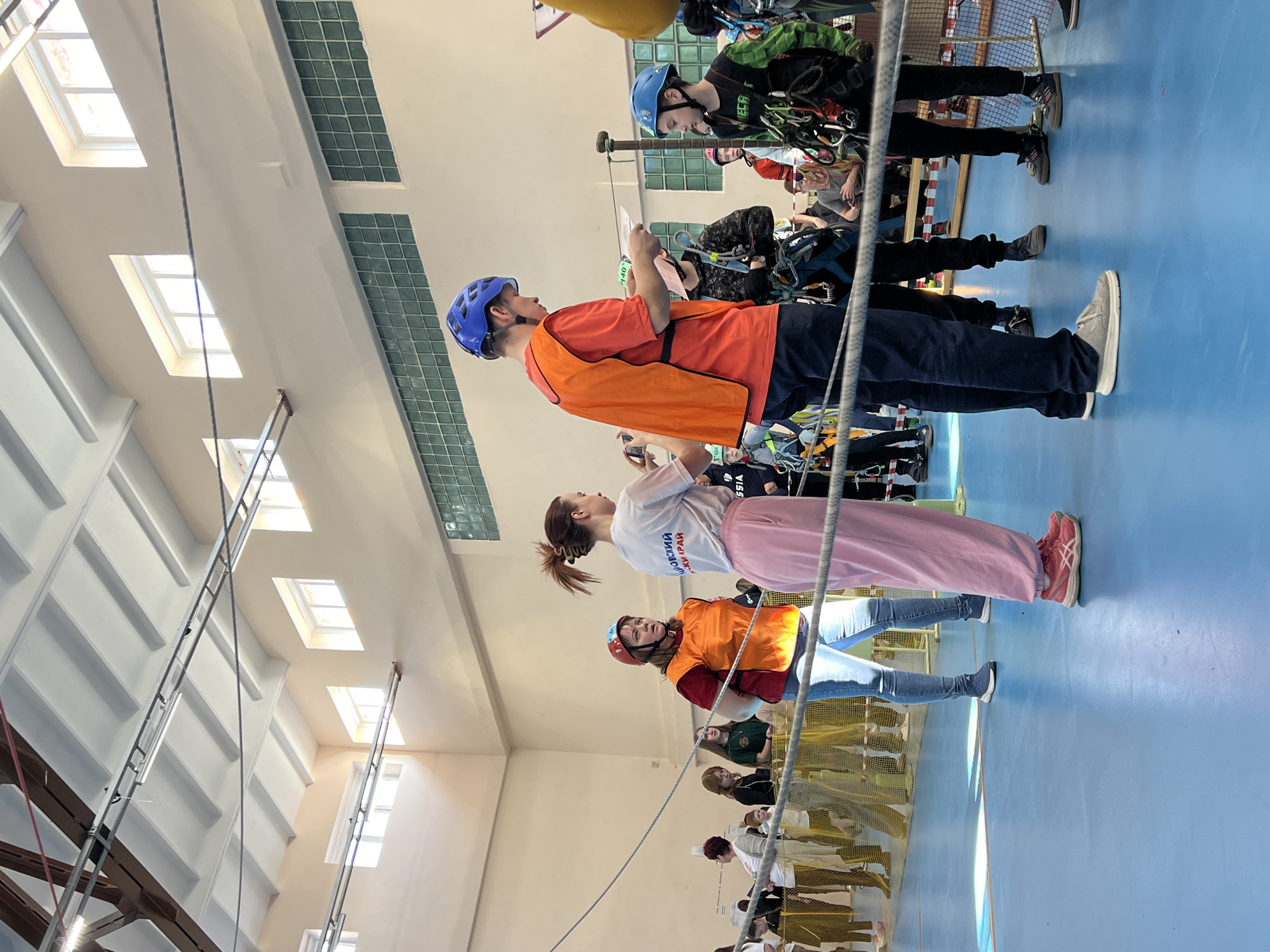 Location: Indoor gymnasium in Berezniki, Russia.
Description: A group of people, mostly young, are engaged in rope climbing activities. Several individuals wear helmets and harnesses, suggesting safety precautions for climbing. A few people appear to be instructors or organizers, possibly reviewing documents or providing instructions. The environment is an indoor sports facility with a blue floor and high ceilings featuring rows of windows for natural light. A climbing rope is visibly rigged, indicating the main activity. The overall scene suggests a training session or competition focused on rope climbing skills.