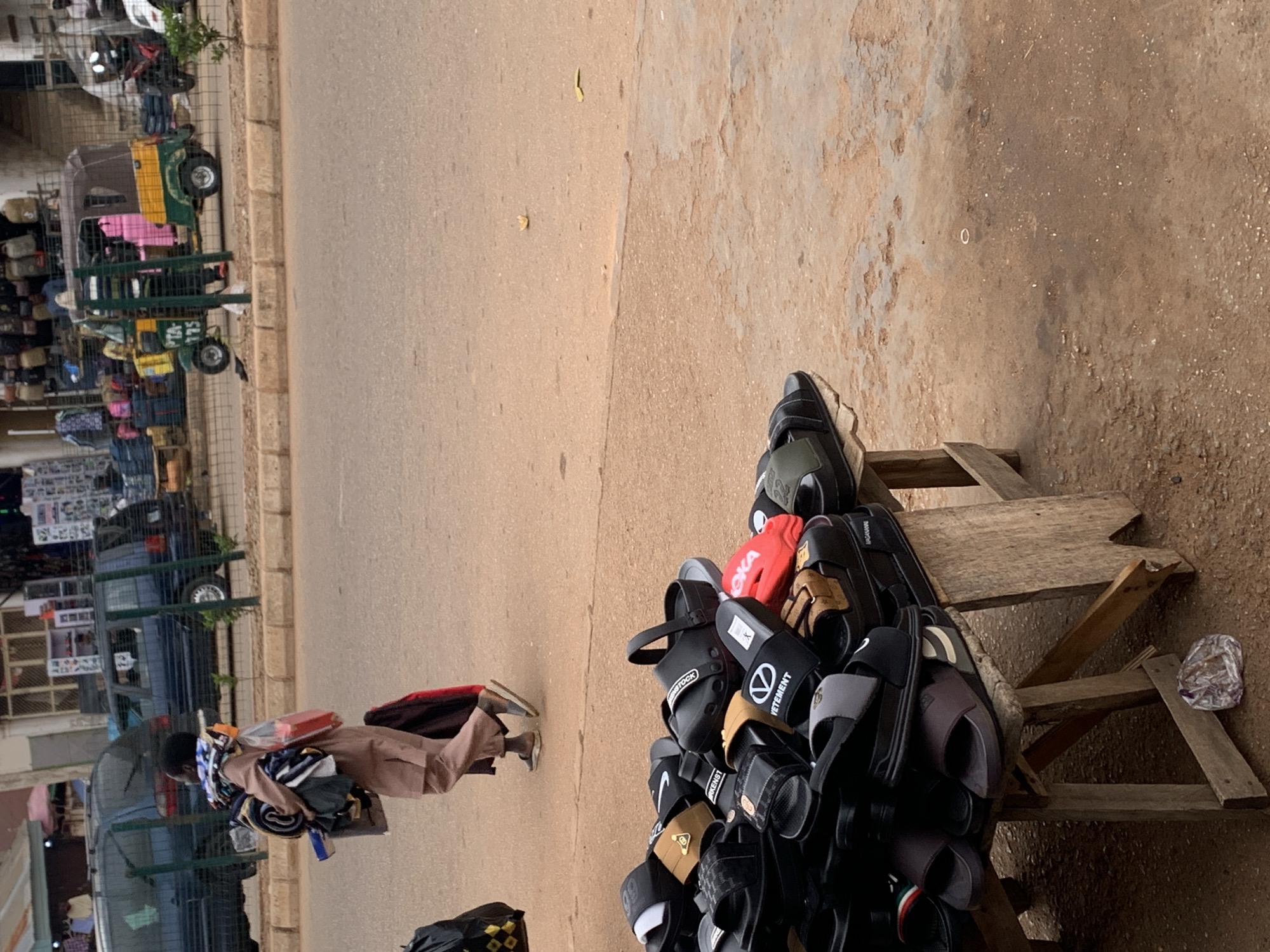 In Bukuru, Nigeria, a man walks along a street carrying a bundle of folded clothing over his shoulder and a red suitcase in his hand. In the foreground, a collection of various sandals and flip-flops is displayed on a wooden table. Behind the man, a yellow and green auto-rickshaw is parked. In the background, other vehicles, stalls with merchandise, and a building are visible. The ground is made of dirt and asphalt, with a curb and a sidewalk bordering the street.