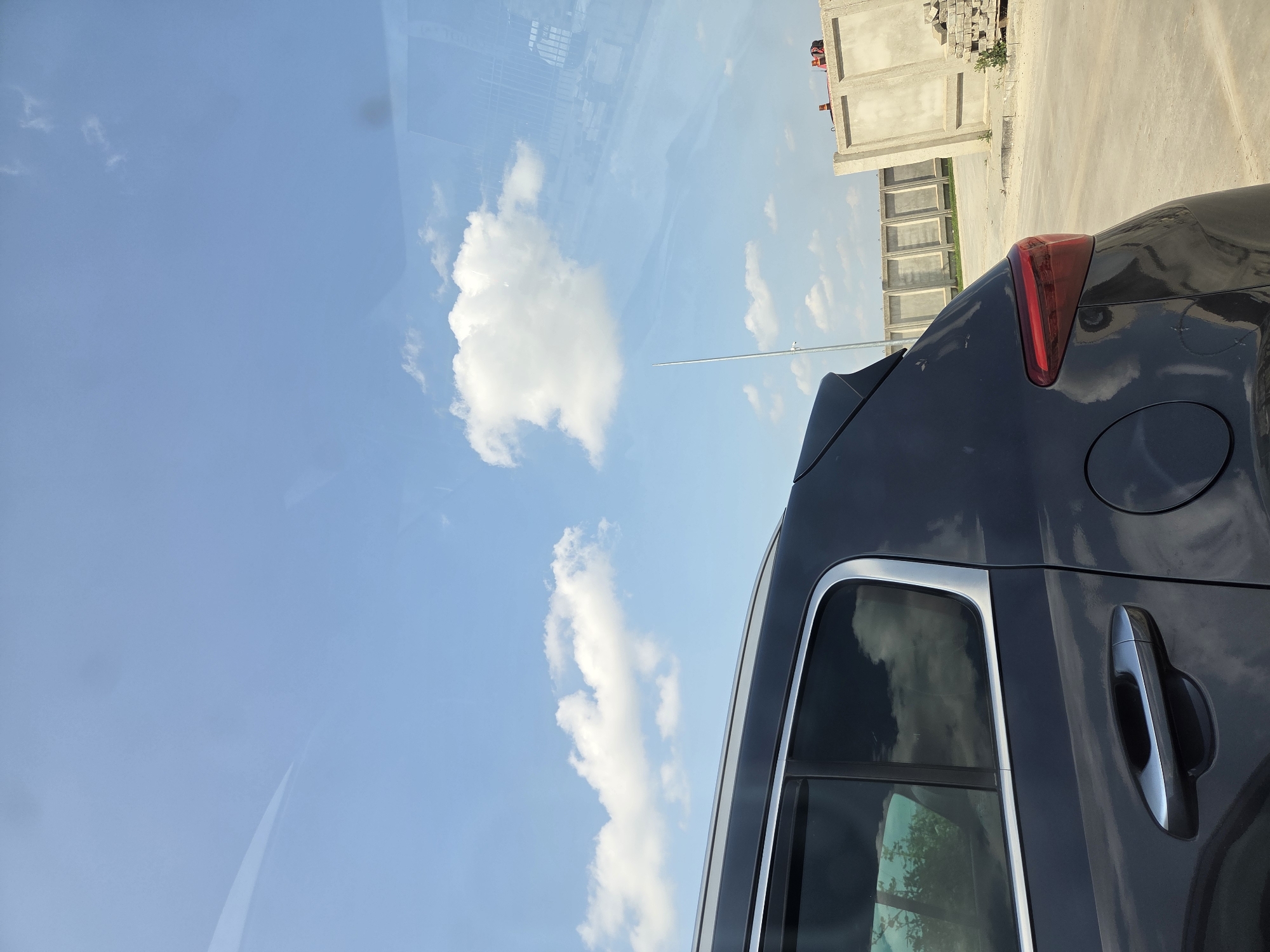 This image, taken in Branipole, Bulgaria, captures the side and rear of a dark-colored car against a bright blue sky with scattered white, fluffy clouds. The original image is vertically oriented, featuring the car on the right side and a significant portion of the sky dominating the left.
The car, possibly an SUV or hatchback, displays a prominent red taillight on its rear passenger side, a circular fuel cap, and a chrome-accented door handle on the rear passenger door. Silver trim outlines the car's windows and runs along the roof, serving as roof rails. Reflections of the sky and some distant foliage are visible in the car's windows and its glossy bodywork.
In the background, beyond the car, are concrete structures resembling large blocks or pre-fabricated walls, suggesting a construction site or industrial area. The ground appears to be concrete or asphalt. A slender, silver-colored pole or antenna extends vertically from the ground near the vehicle.
The scene is set outdoors under clear, sunny weather, indicating midday or early afternoon. No specific activity is depicted; the car appears to be parked. There is no visible text in the image.