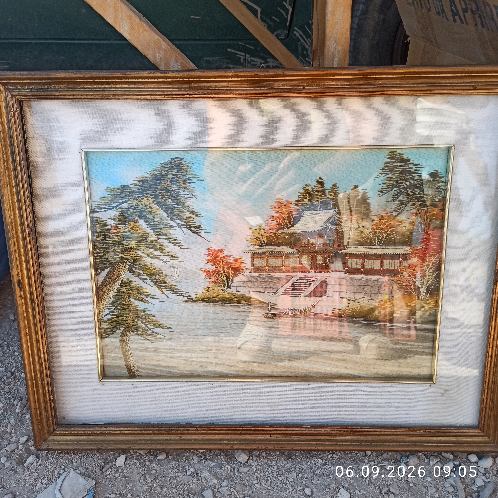 A framed artwork depicting a traditional Japanese landscape is displayed outdoors. The artwork, encased in a weathered golden frame, showcases a serene scene with a Japanese-style building with multiple levels and an ornate roof, surrounded by trees displaying autumn foliage. A body of water, possibly a lake or river, is in the foreground, with a small boat gently floating on its surface. A large pine tree with intricate branches dominates the left side of the composition. The artwork appears to be an embroidery or a similar textile art form, given the texture and fine detail visible. Reflections on the glass covering the artwork indicate the presence of overhead light and possibly a person taking the photograph. The background of the overall image reveals a dark green surface and wooden elements, suggesting it is propped up or leaning against something. The ground is covered in gravel and small stones, typical of an outdoor setting. A timestamp in the lower right corner indicates "06.09.2026 09:05." The artwork is located in Sannicandro di Bari, Italy.
