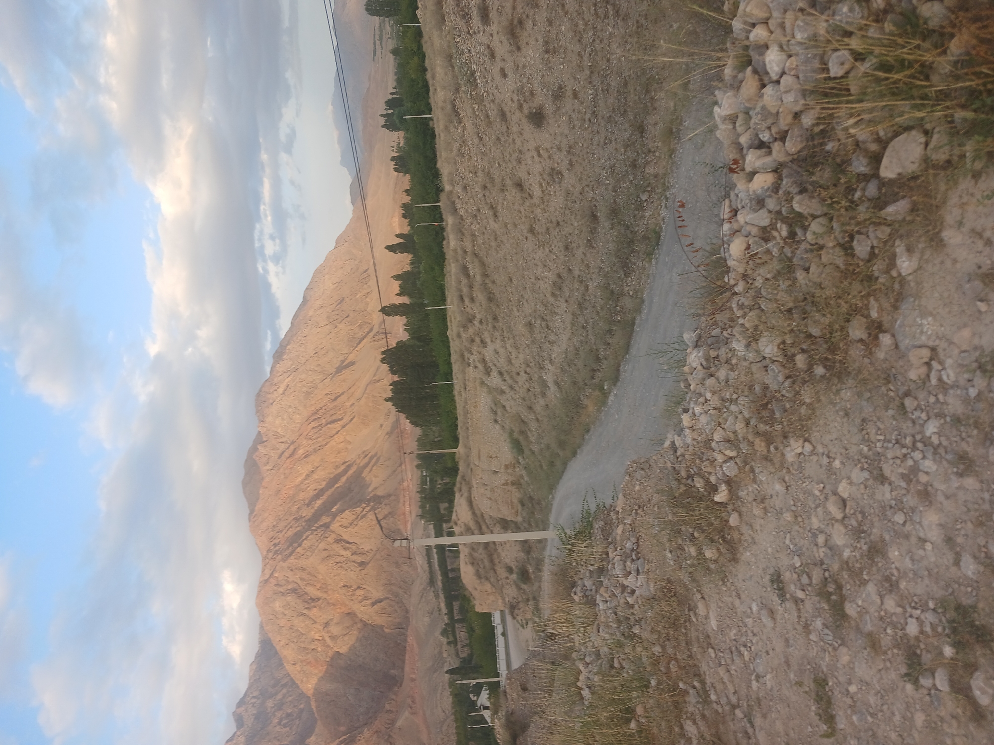 This is an outdoor, landscape photograph taken in the city of Aksai, Kyrgyzstan. The image depicts a rural scene with mountains in the background and a dirt road winding through the foreground. The mountains are rugged and appear to be illuminated by the golden light of either sunrise or sunset. A valley with scattered trees and vegetation lies between the mountains and the road. Several utility poles with wires are visible, suggesting the presence of infrastructure in the area. The ground in the foreground is covered with gravel, rocks, and dry grass. The sky is partly cloudy with blue patches visible. There are no people or animals present in the image.