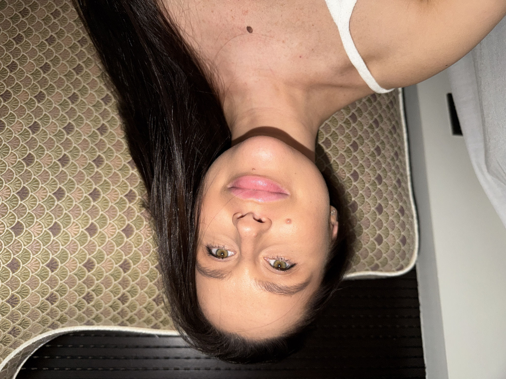 A woman with dark hair is lying on a pillow with a scalloped pattern, indoors. The woman is fair-skinned with light-colored eyes. She is wearing a white top. The location is not discernible from the image; therefore, there is no evidence to suggest it is Khimki, Russia.
