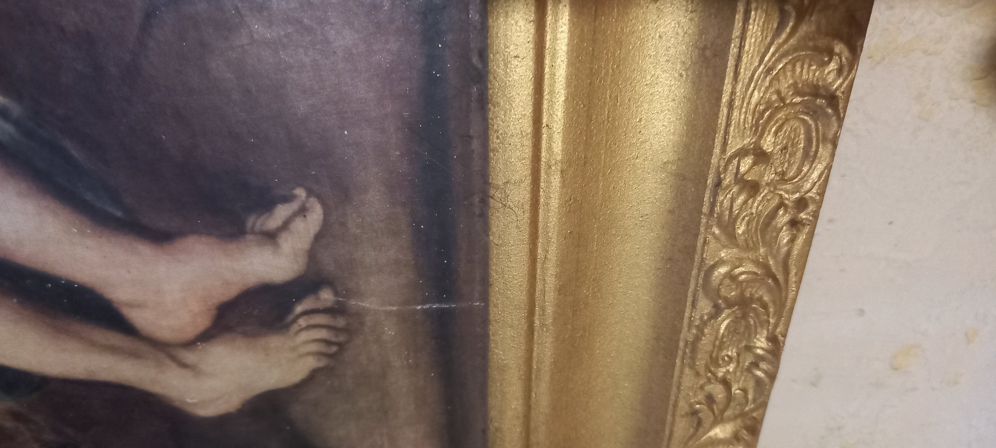 The image shows a close-up view of a painting's lower section, specifically focusing on the feet of a figure and a decorative gold frame. The feet appear to be rendered in a realistic, classical style, with visible toes and ankles. The painting's background is dark and textured, suggesting a somber or dramatic scene. The frame is ornate, with detailed carvings and a gilded finish. The image is taken from an angle that emphasizes the texture and dimensionality of both the painting and the frame. The location in Kryvyi Rih, Ukraine, is not discernible from the image content.