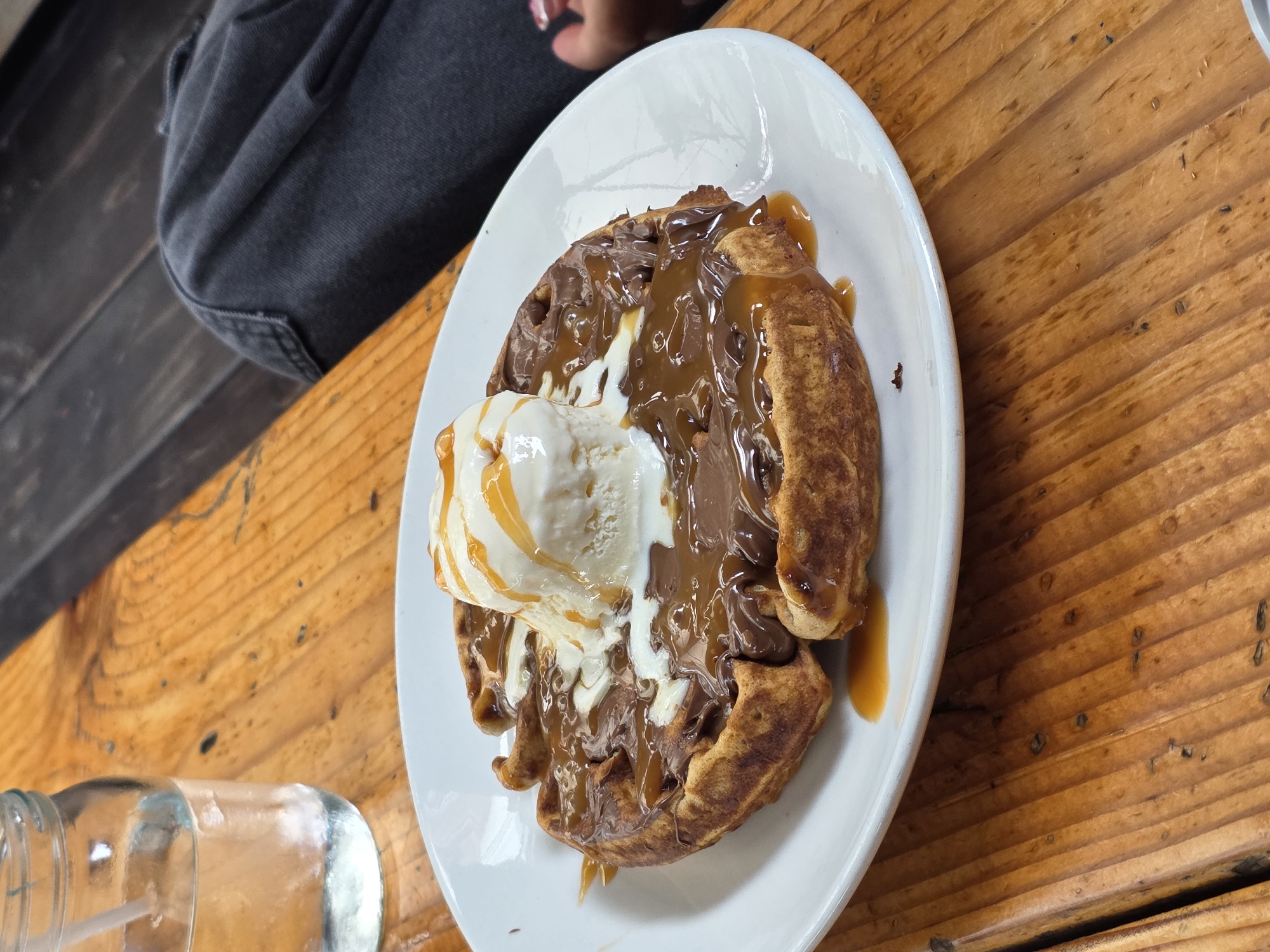 The image depicts a waffle dessert on a white plate, topped with ice cream, chocolate, and caramel sauce. The plate sits on a wooden table. In the background, a portion of a person's clothing is visible, along with a glass filled with water. The scene appears to be in a restaurant or cafe, possibly in Ensenada, Mexico, but this location cannot be confirmed based solely on the image.