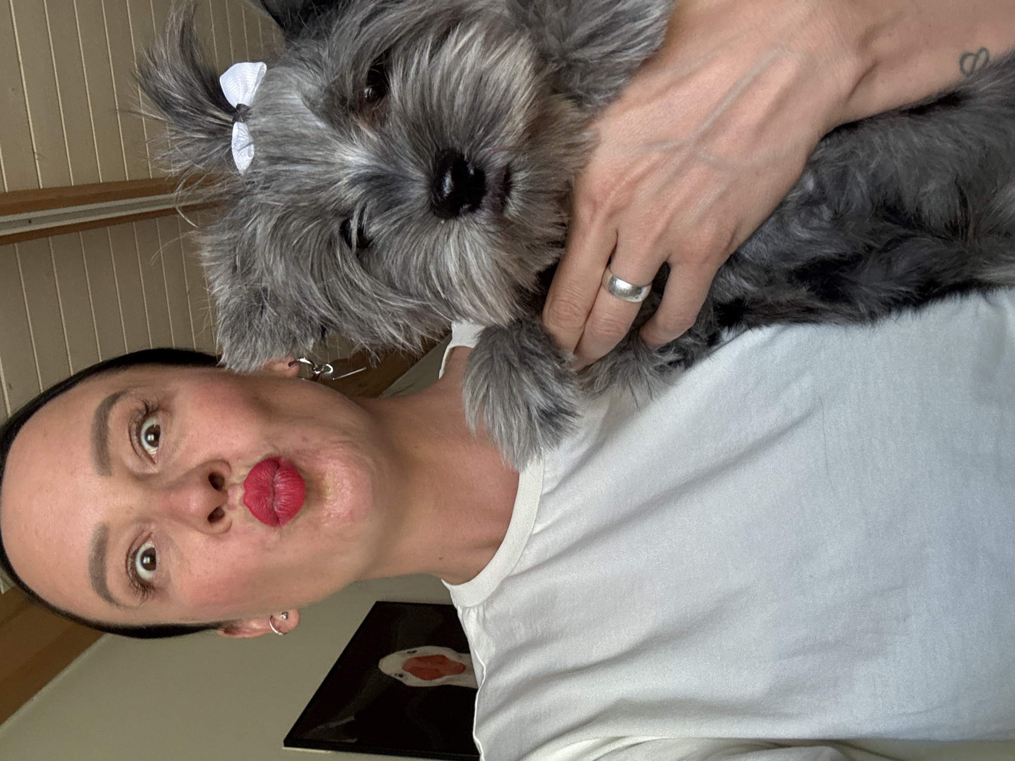 A woman with dark hair pulled back is making a kissing face, holding a gray dog with a white bow on its head. The woman is wearing a white t-shirt and has silver earrings and a ring on her finger. The background includes vertical wooden paneling, and part of a framed picture with a white and red object is visible. The location is not discernible from the image.